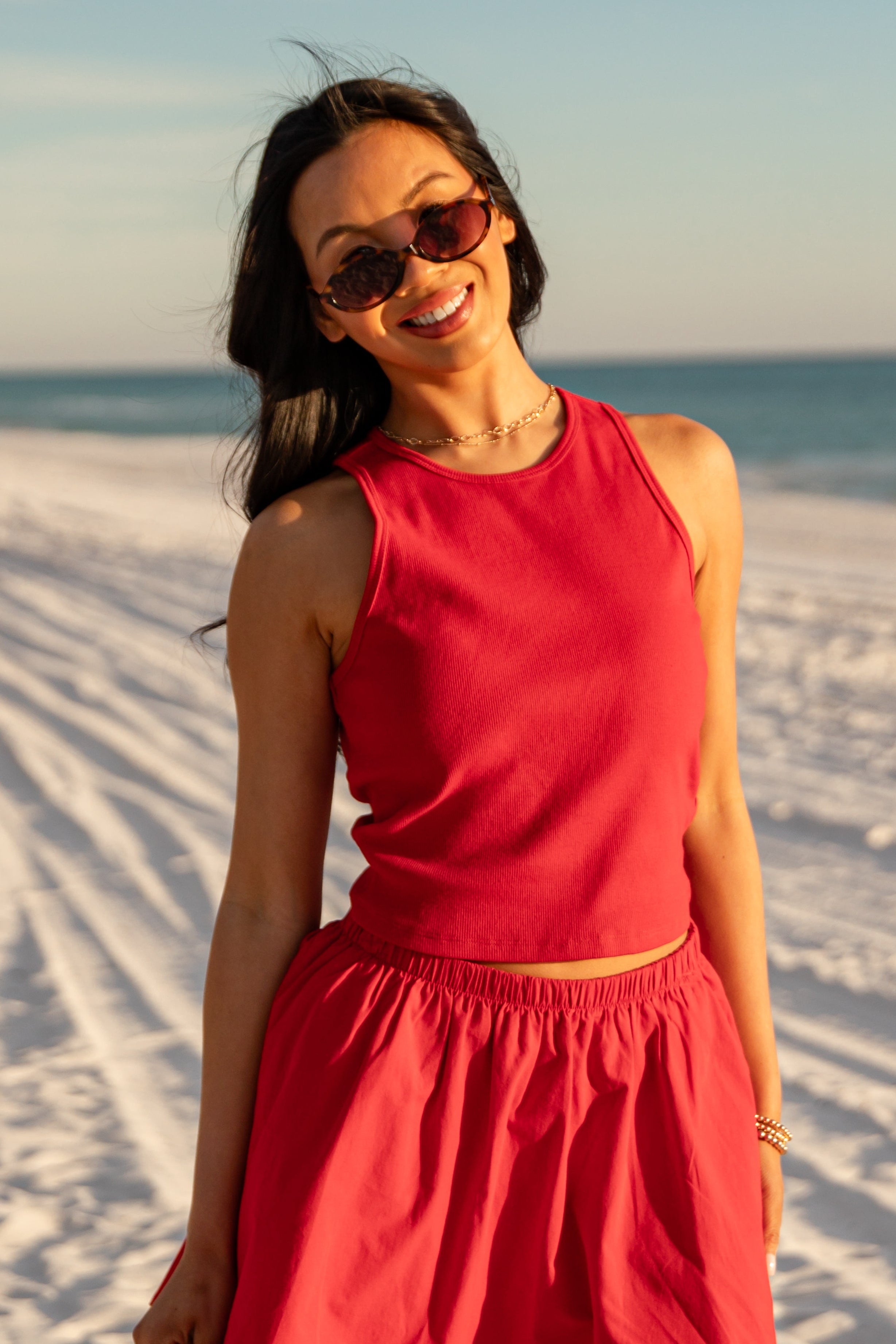 Woman in a red outfit standing on a sandy beach with sunglasses.