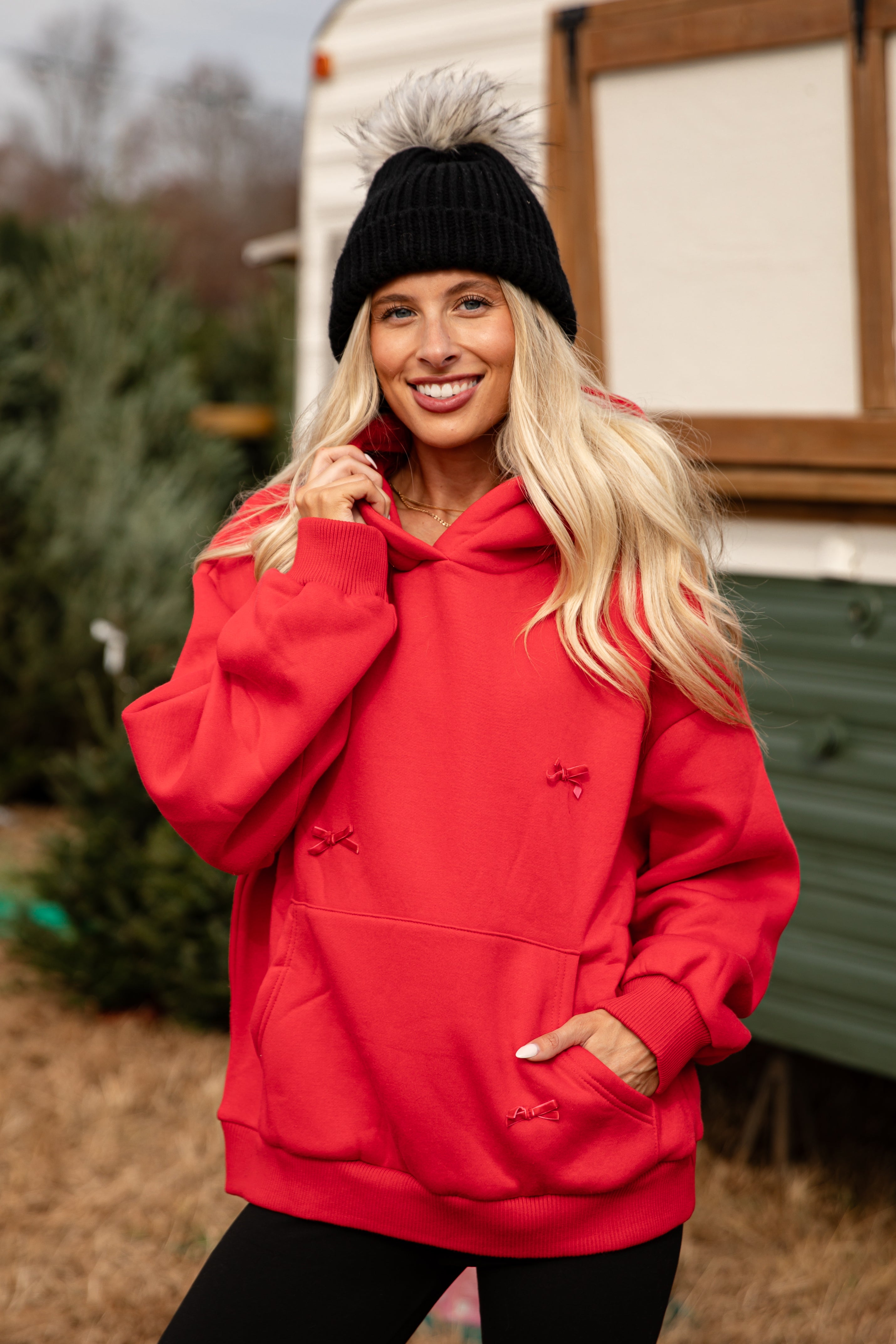 Woman wearing a red hoodie with a logo, standing outdoors near trees and a trailer.