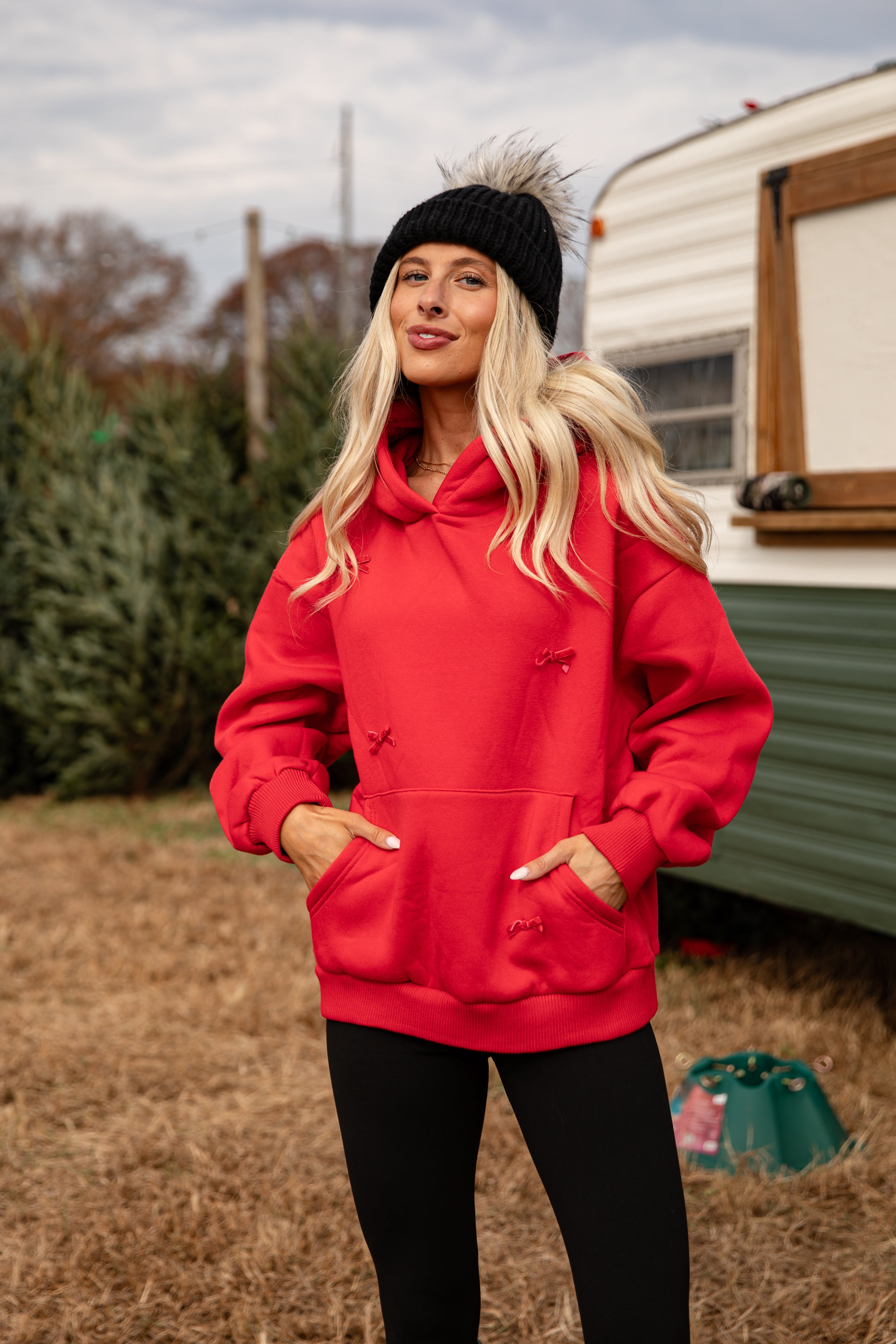 Woman wearing a red hoodie and black beanie standing outdoors with a trailer and trees in the background.
