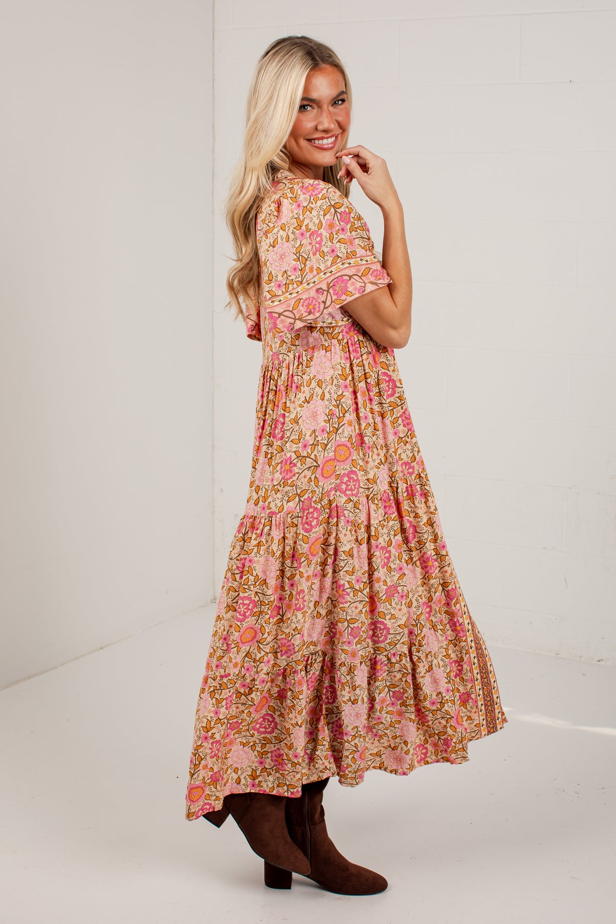 A woman modeling a long tiered maxi dress with a pink, orange, and yellow floral print, paired with brown ankle boots, photographed against a plain white studio background.