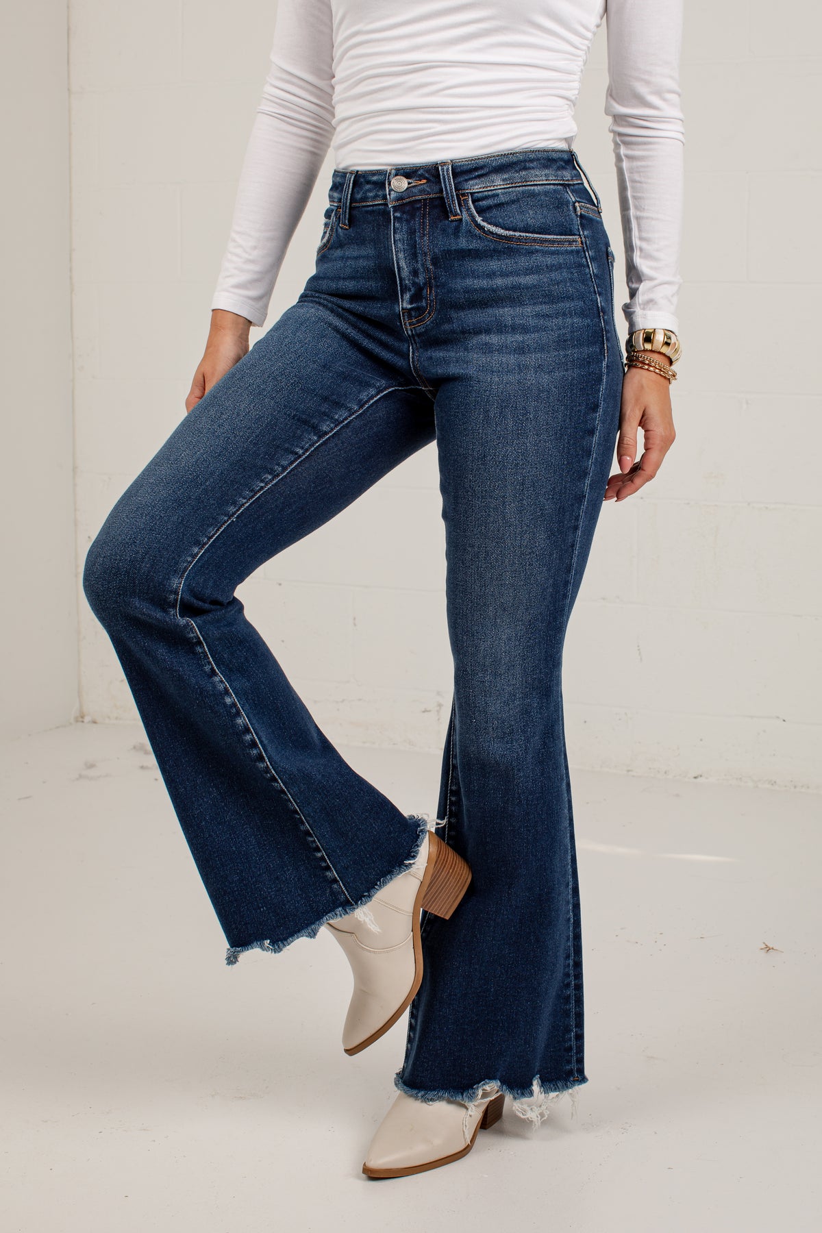 A model wearing dark blue flare jeans with a raw frayed hem, styled with a white long-sleeve top and white cowboy boots against a white studio background.