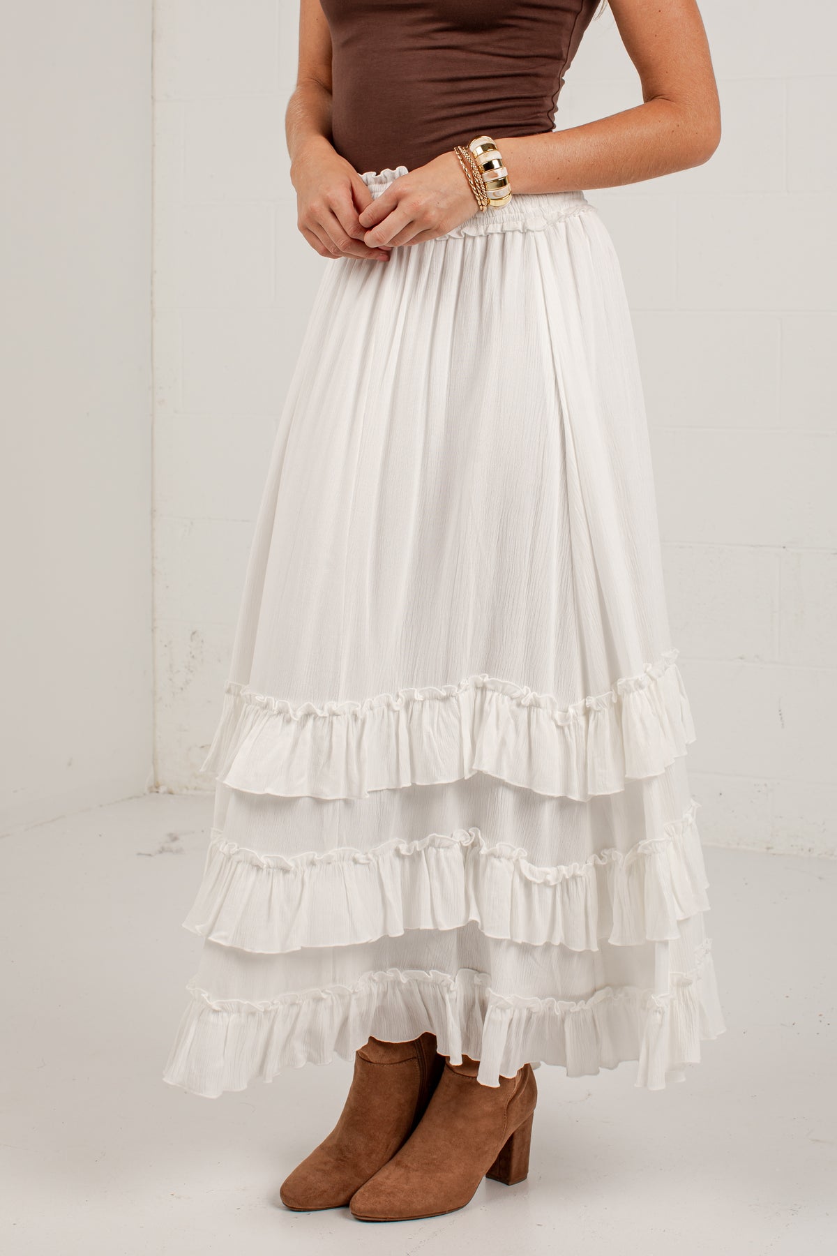 A model wearing a long white tiered maxi skirt with ruffle trim, paired with a brown top and brown suede boots, standing against a white wall.