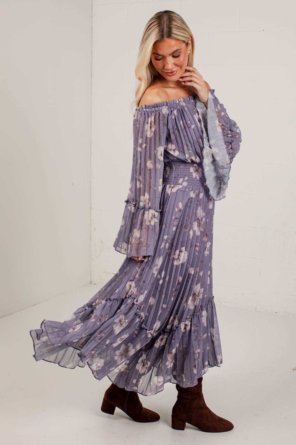A woman modeling a lavender off-the-shoulder maxi dress with a white floral print, tiered ruffle skirt, and long bell sleeves, paired with brown ankle boots in a bright studio setting.
