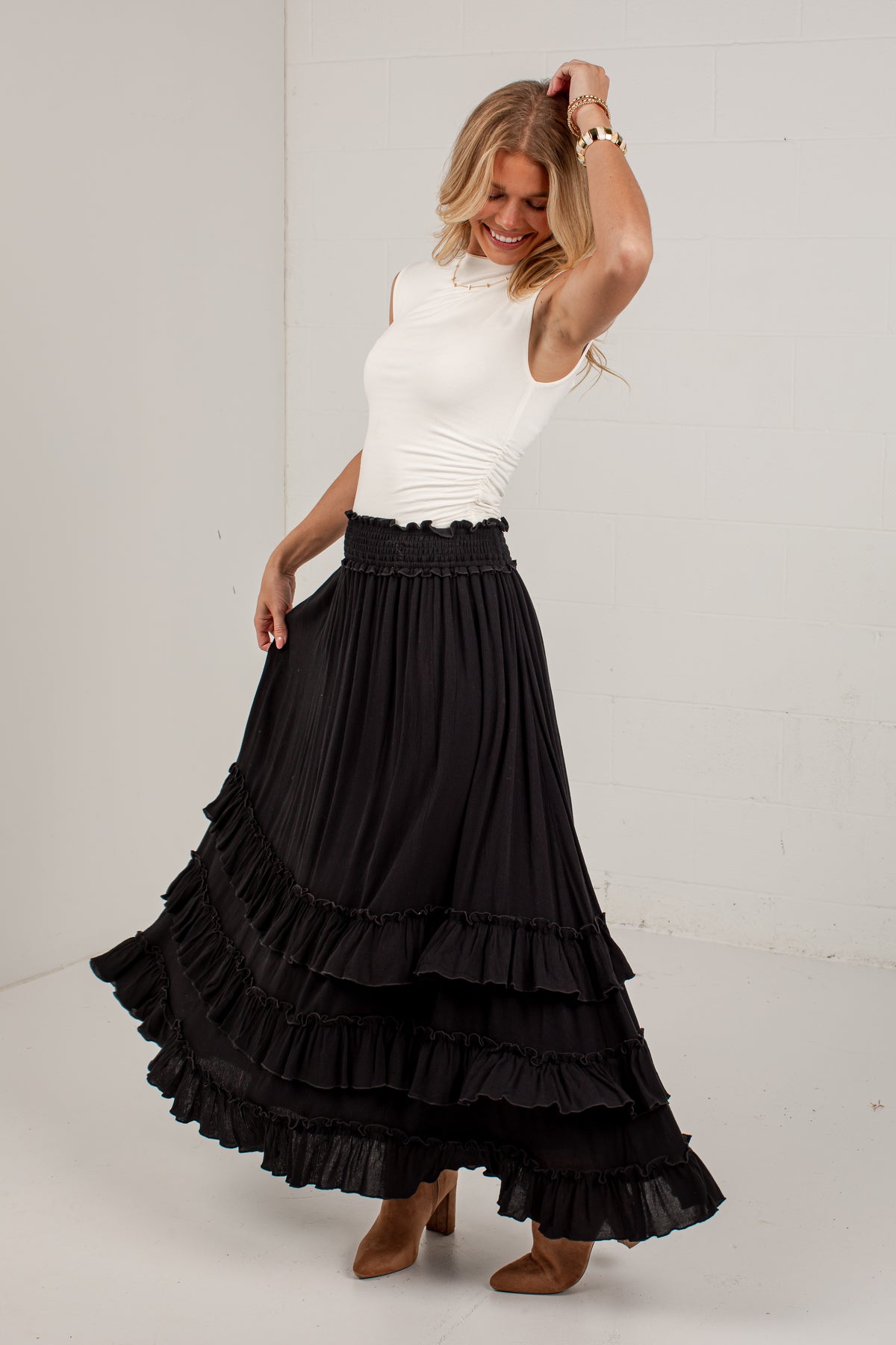 A woman modeling a long black tiered maxi skirt with a smocked waistband, paired with a white sleeveless top and brown heeled boots, standing against a white studio wall.