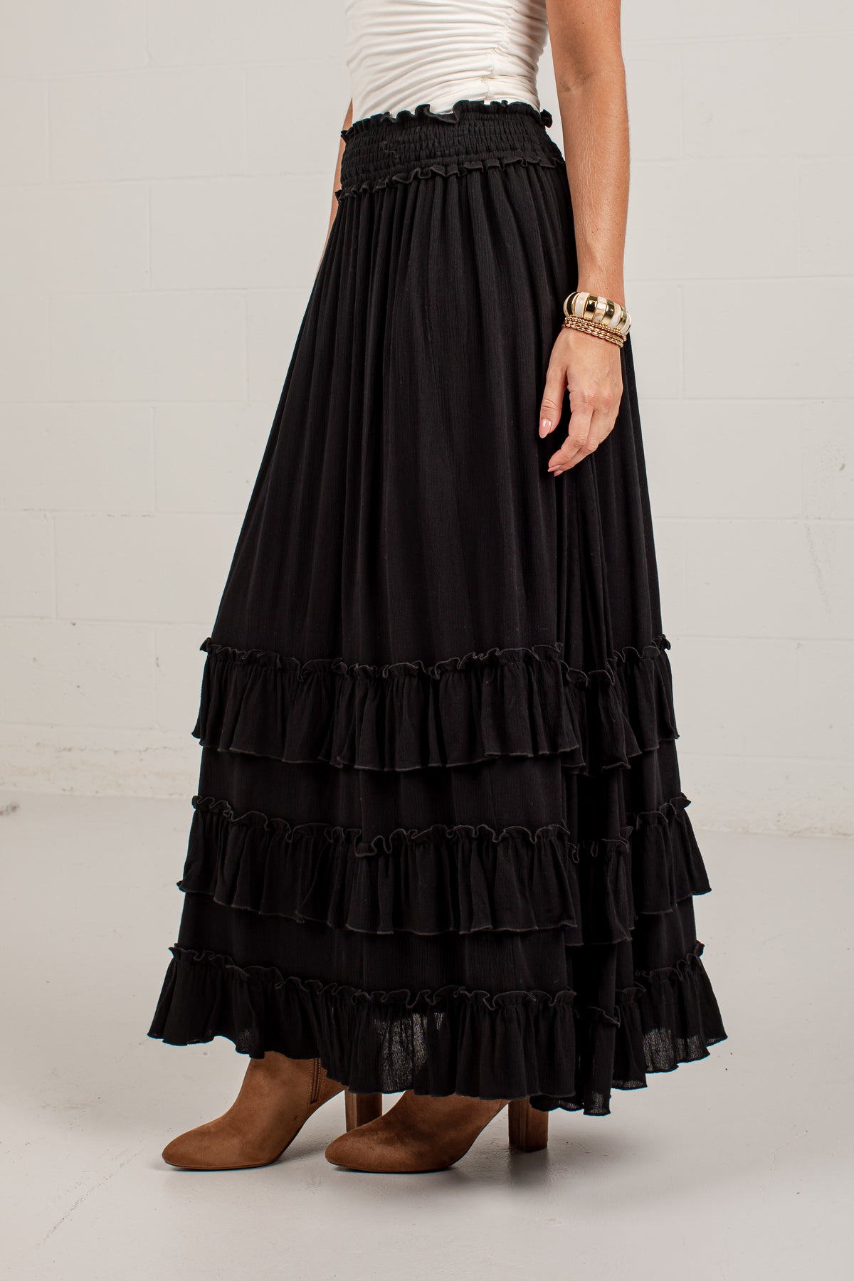 A black tiered maxi skirt with a smocked elastic waistband, shown on a model wearing a white top and brown boots against a white studio wall.