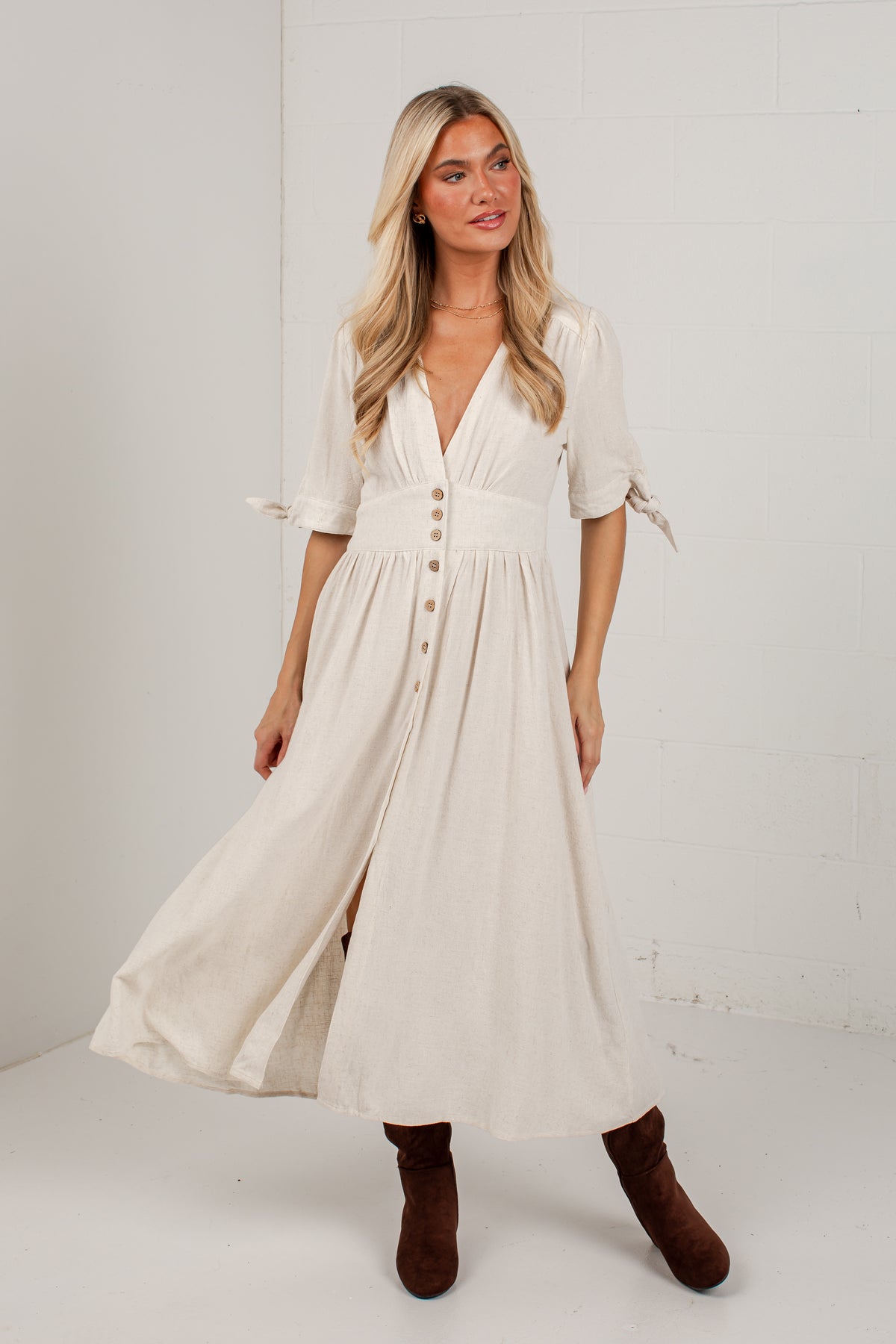 A woman modeling a cream-colored midi dress with short sleeves, a V-neckline, and a button-down front, paired with brown suede boots, set against a white studio background.