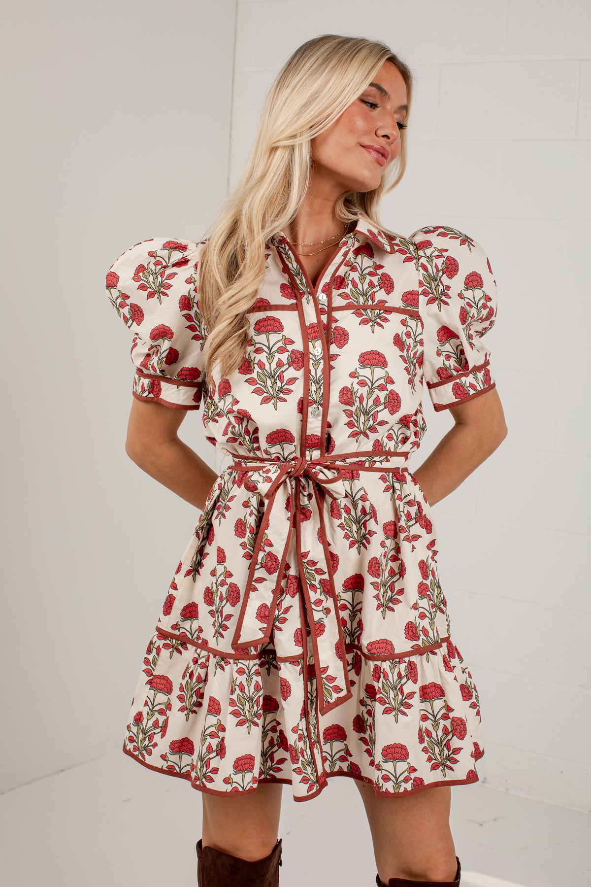 A woman modeling a short puff-sleeve dress with a red and green floral print on a cream background, featuring a tie waist belt and tiered skirt, set against a plain white studio background.
