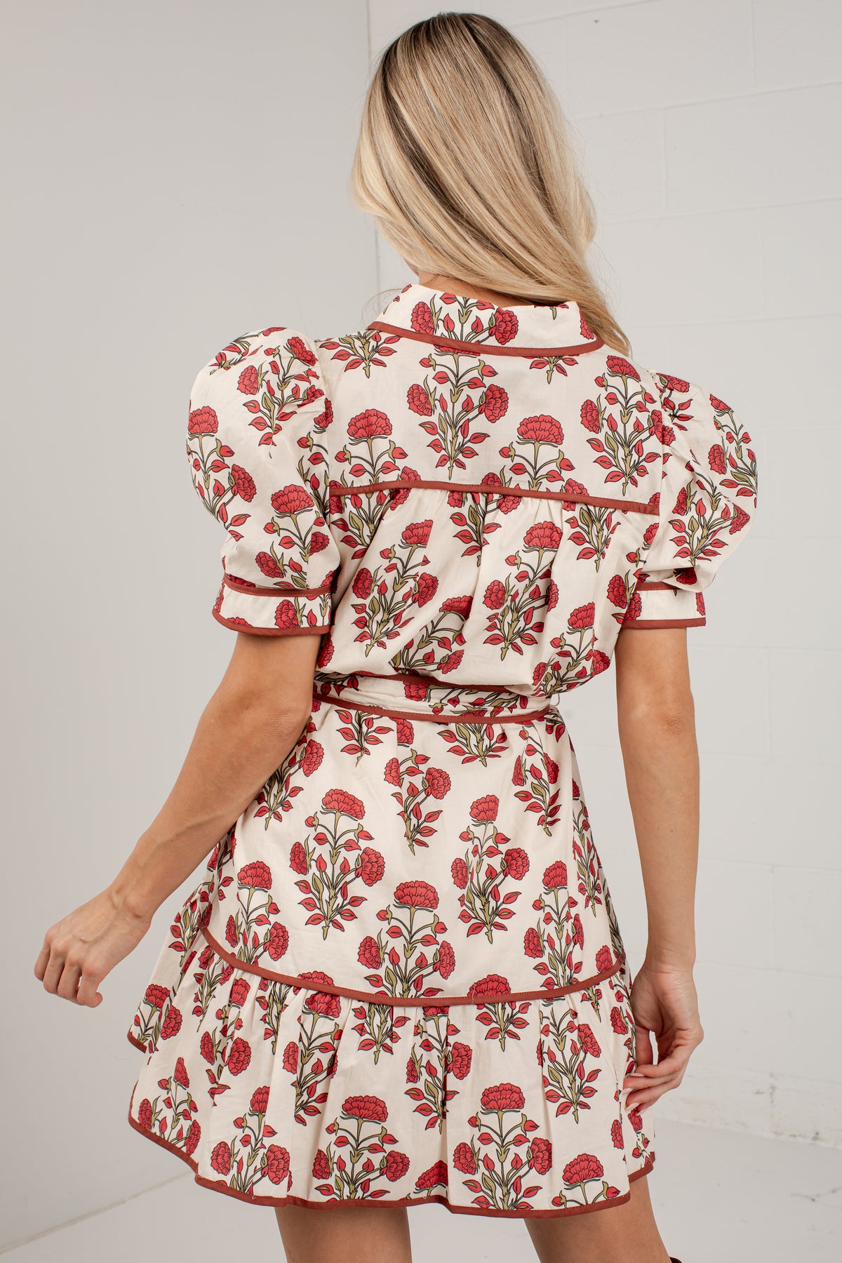 A woman shown from the back wearing a cream-colored mini dress with a red and green floral pattern, puffed short sleeves, and a ruffled waist.