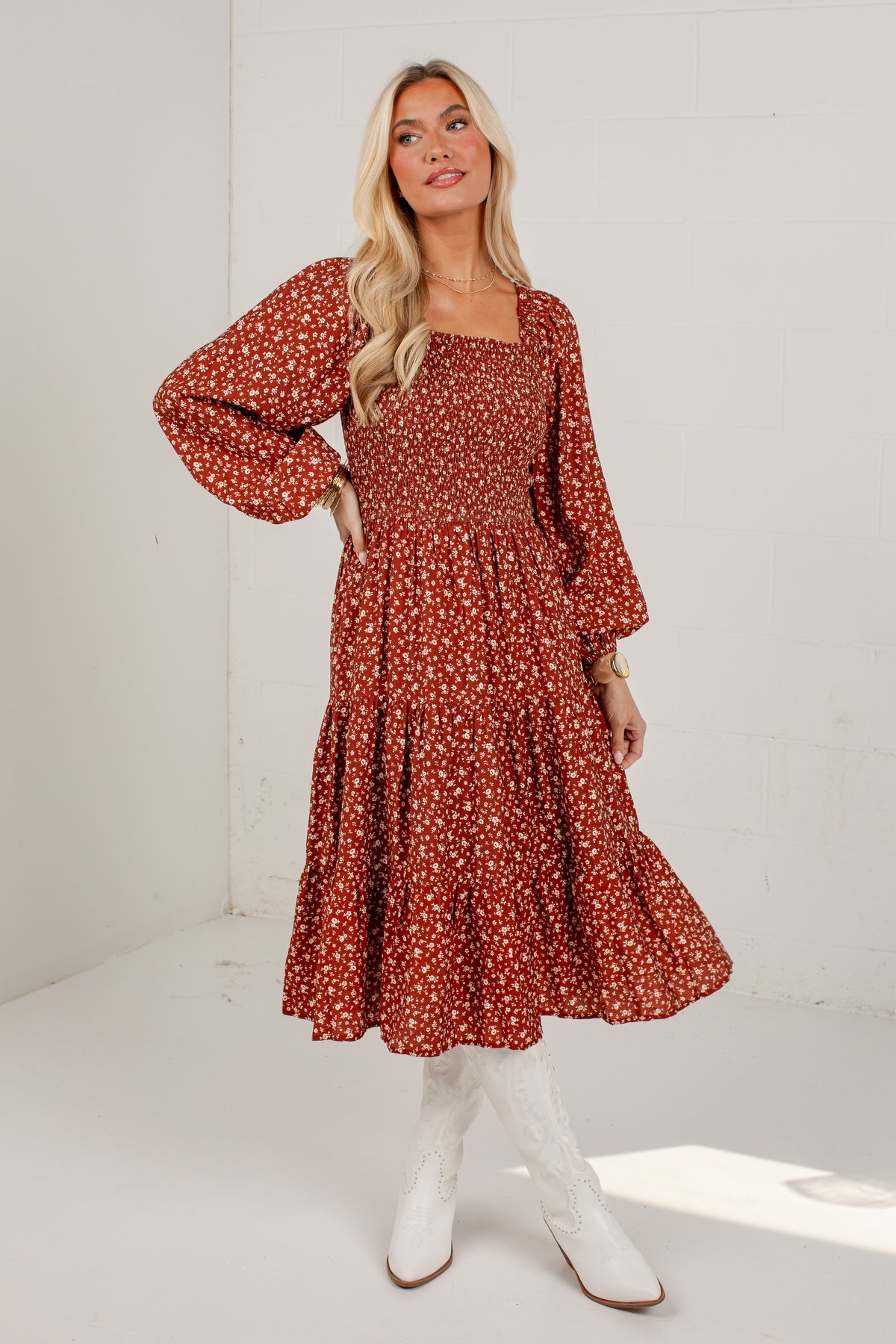 A woman modeling a rust-red midi dress with a white floral pattern, featuring a square neckline, long balloon sleeves, and a tiered skirt, paired with white cowboy boots against a white studio background.