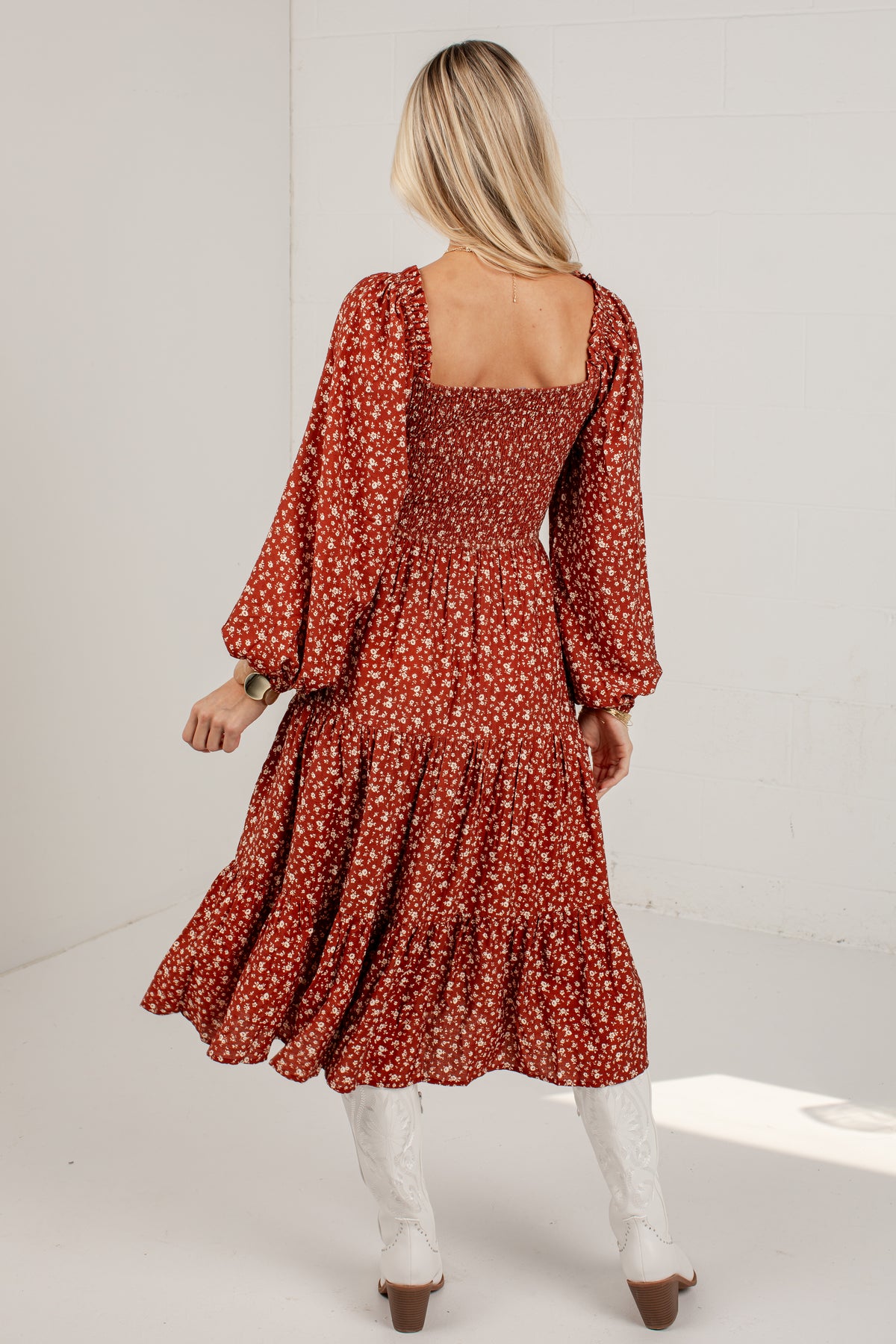 A woman shown from the back wearing a rust-red midi dress with a white floral pattern, long balloon sleeves, and a smocked bodice. She is also wearing white cowboy boots against a white studio background.