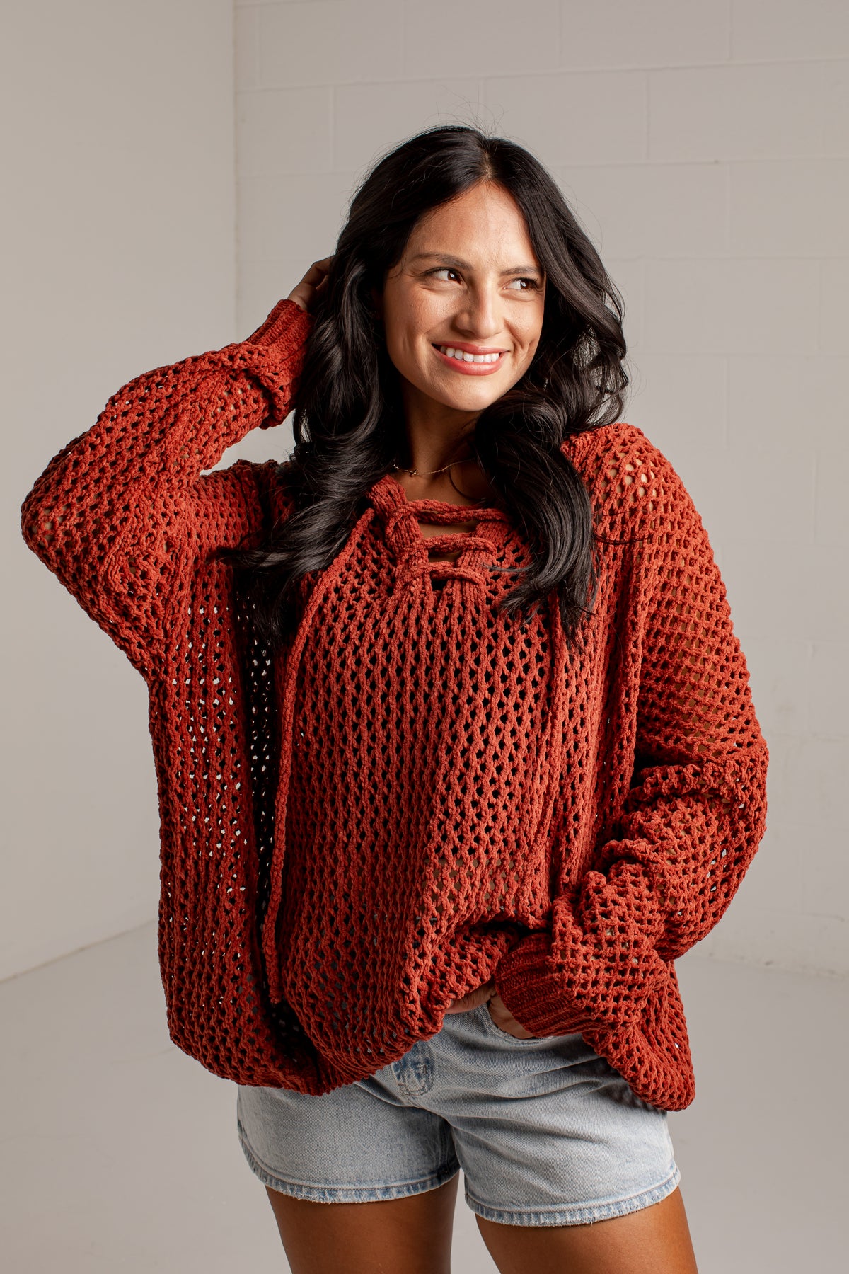A woman wearing a rust-orange chunky knit crochet sweater with a tie-neck detail, paired with light-wash denim shorts, posing against a light gray brick wall.