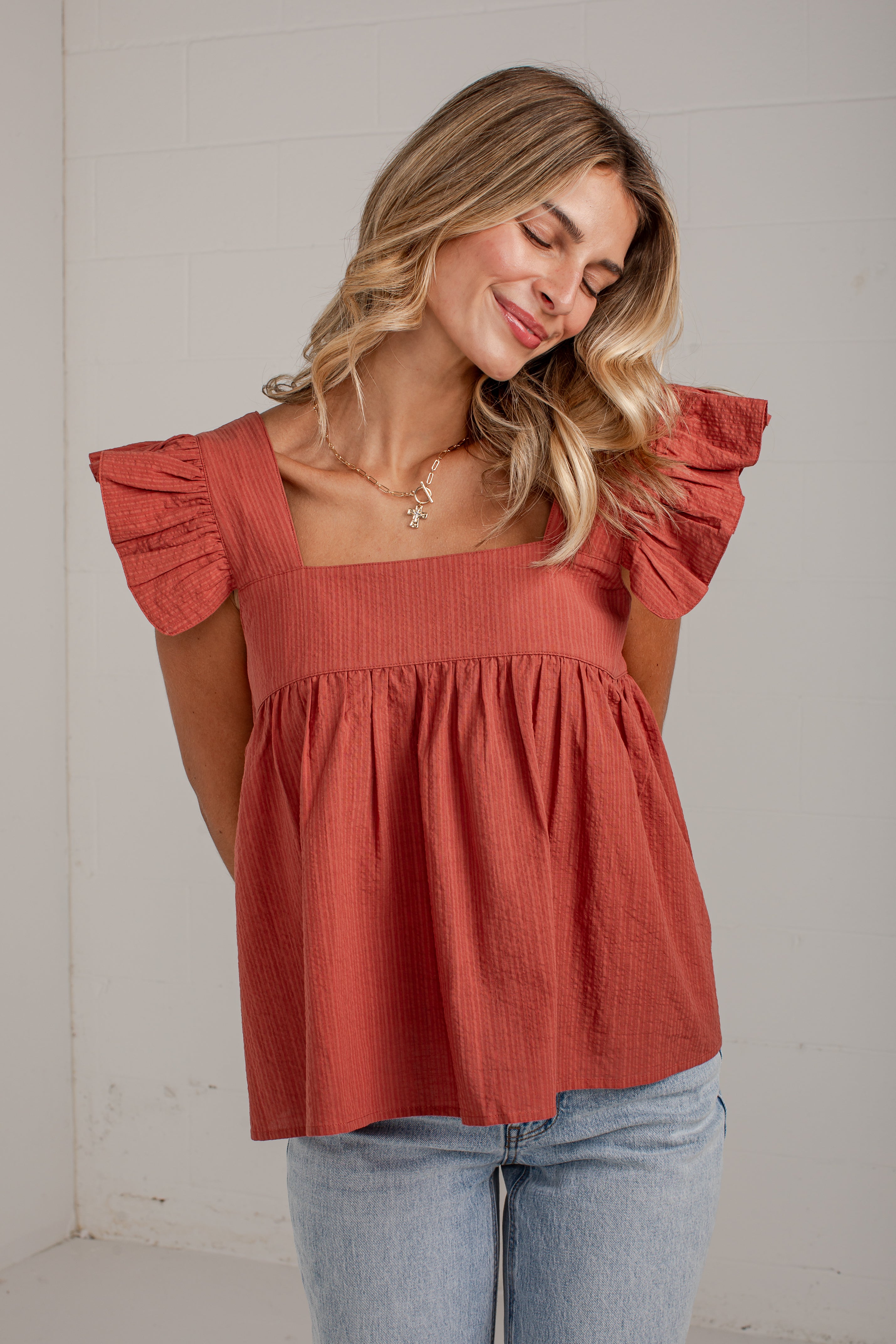 A woman wearing a terracotta-colored crinkle-textured top with ruffled sleeves and a square neckline, paired with light-wash blue jeans, standing against a white paneled wall.