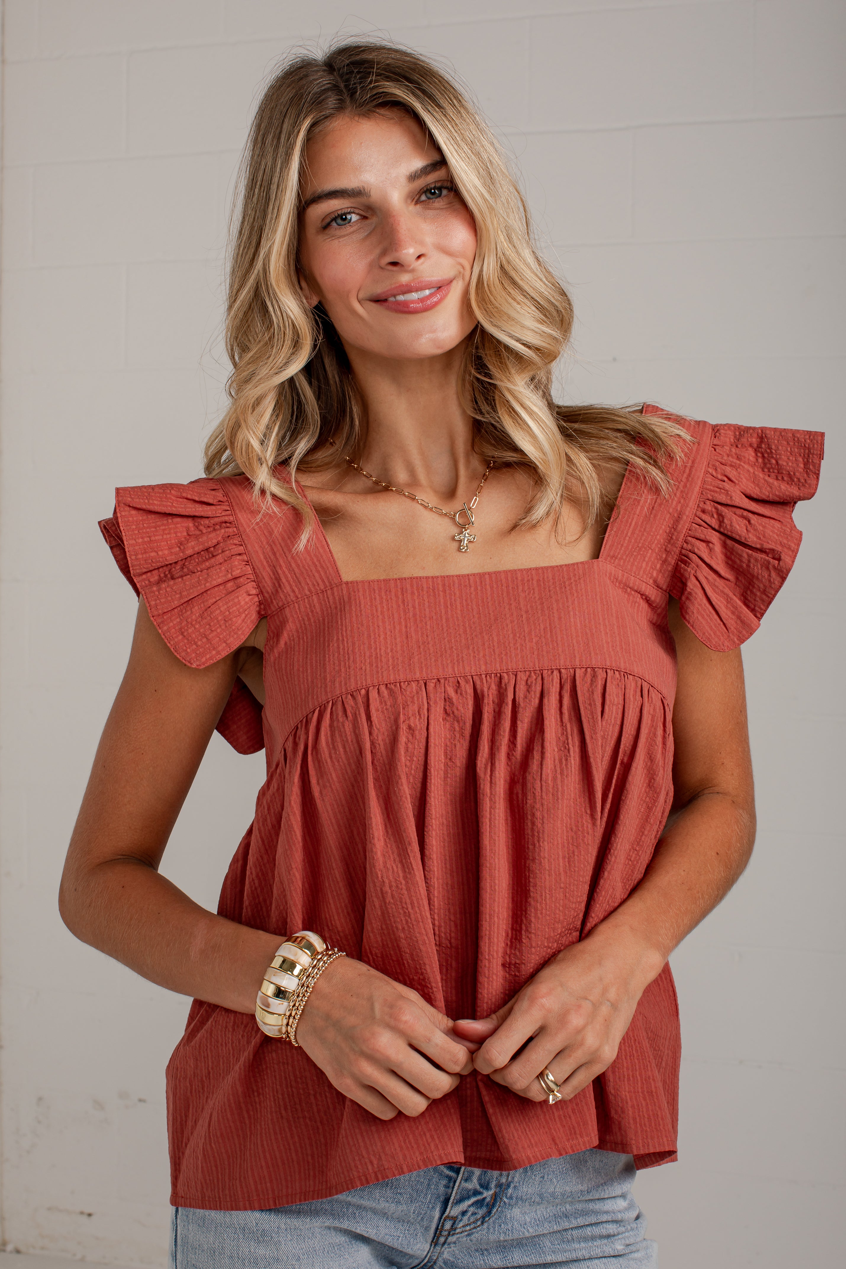 A woman wearing a terracotta-colored ruffle-sleeve top with a square neckline, paired with light blue jeans, standing against a white paneled wall.