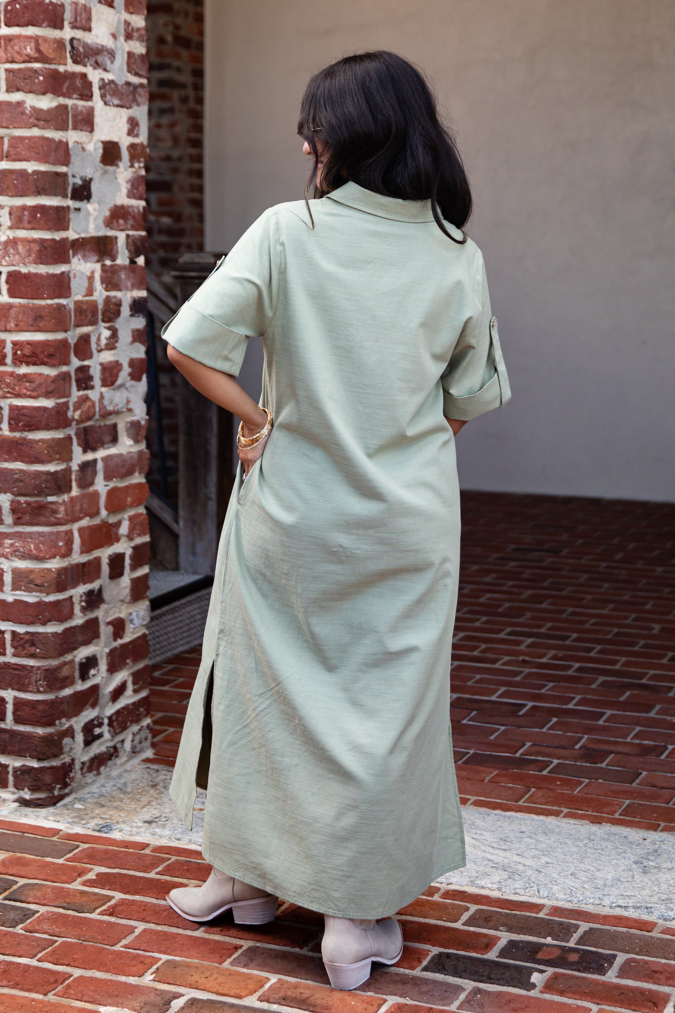 A woman viewed from behind wearing a light sage green linen shirt dress in an outdoor setting with a brick wall and red brick floor.