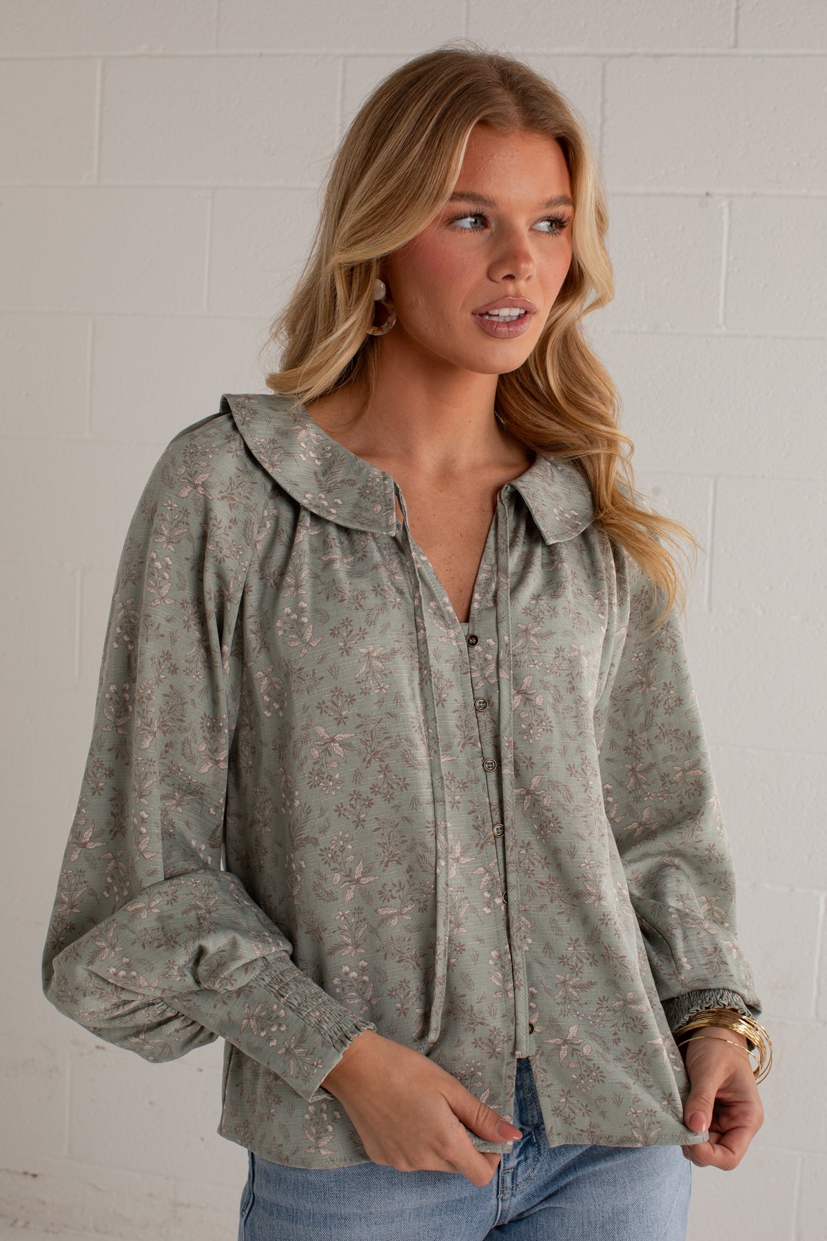 A woman wearing a sage green long-sleeve blouse with a delicate floral pattern, paired with light blue jeans, standing against a white brick wall.