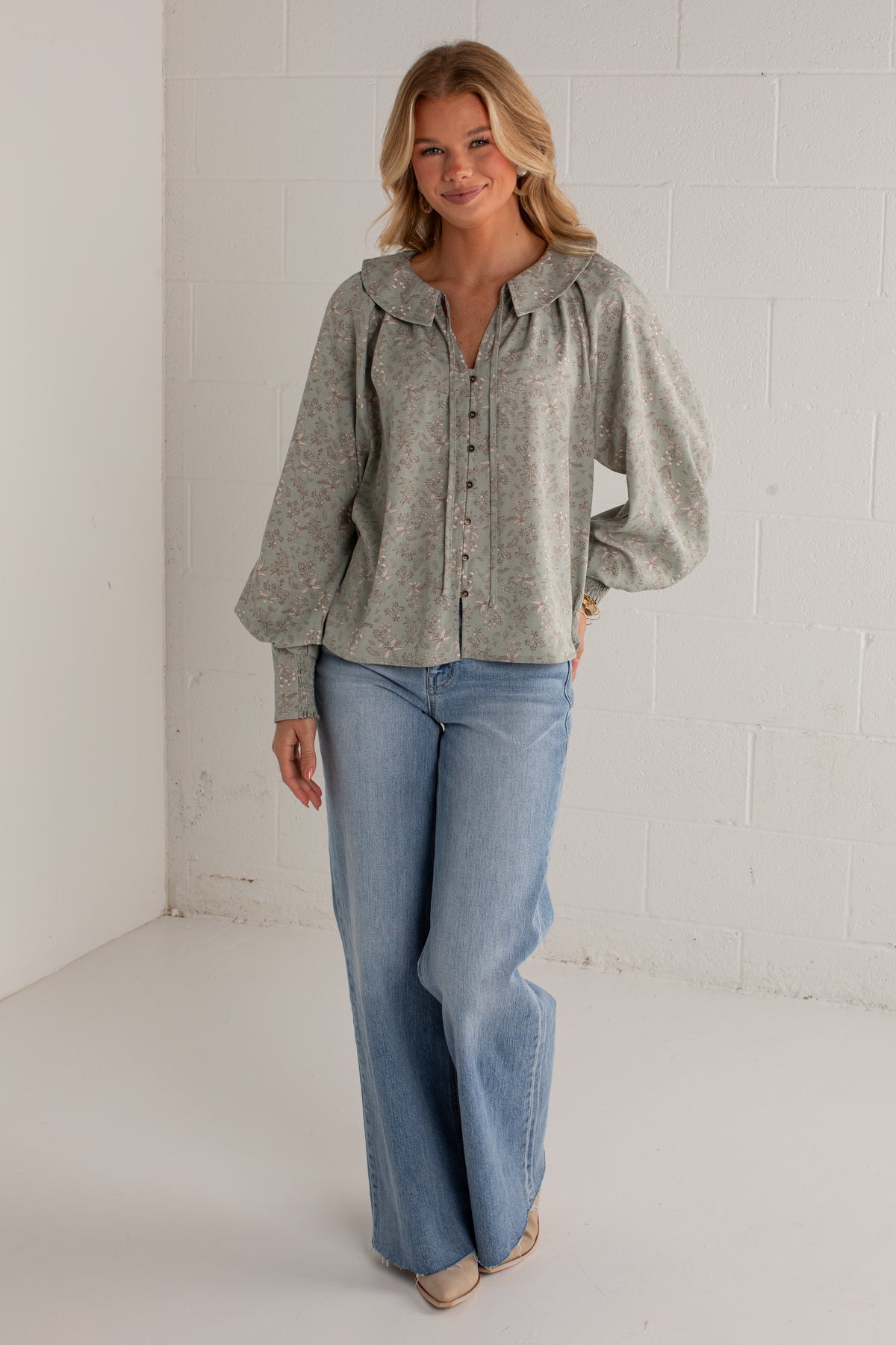 A woman modeling a light sage green long-sleeve button-up blouse with a wide collar and voluminous sleeves, paired with light-wash wide-leg jeans, standing against a white brick wall.