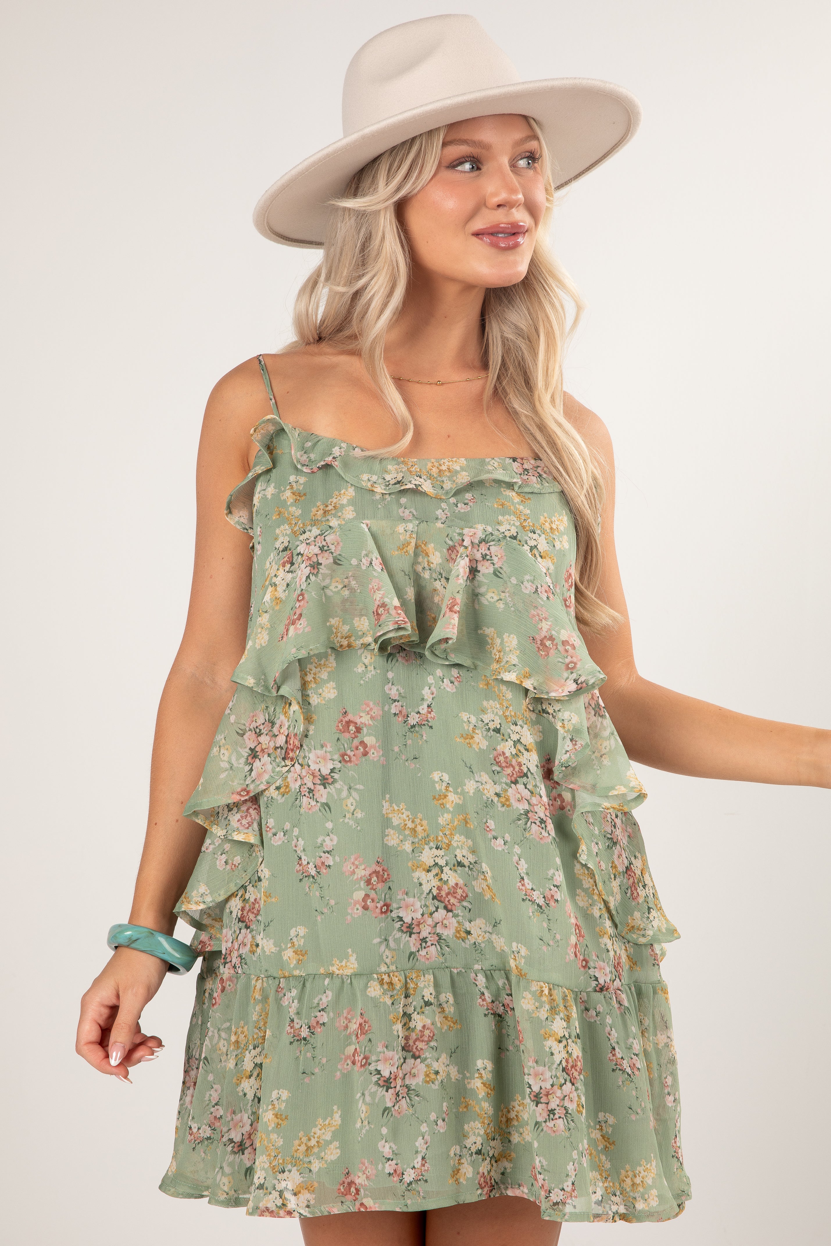 A woman wearing a sage green tiered ruffle mini dress with a pink and yellow floral print, styled with a beige wide-brim hat and a turquoise bracelet against a light gray studio background.