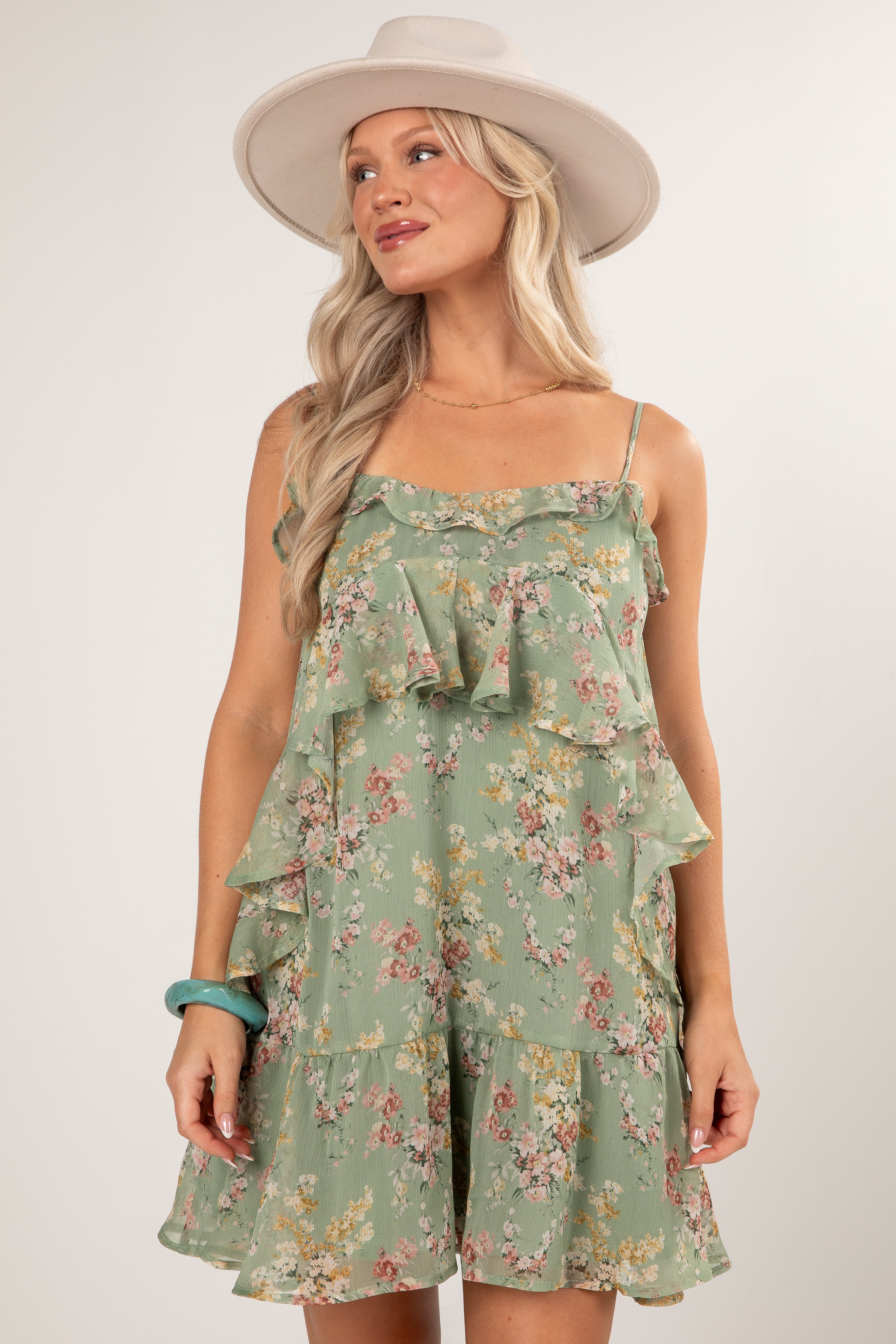 A woman wearing a sage green ruffled spaghetti-strap romper with a pink and yellow floral print, styled with a wide-brimmed cream hat against a light gray studio background.