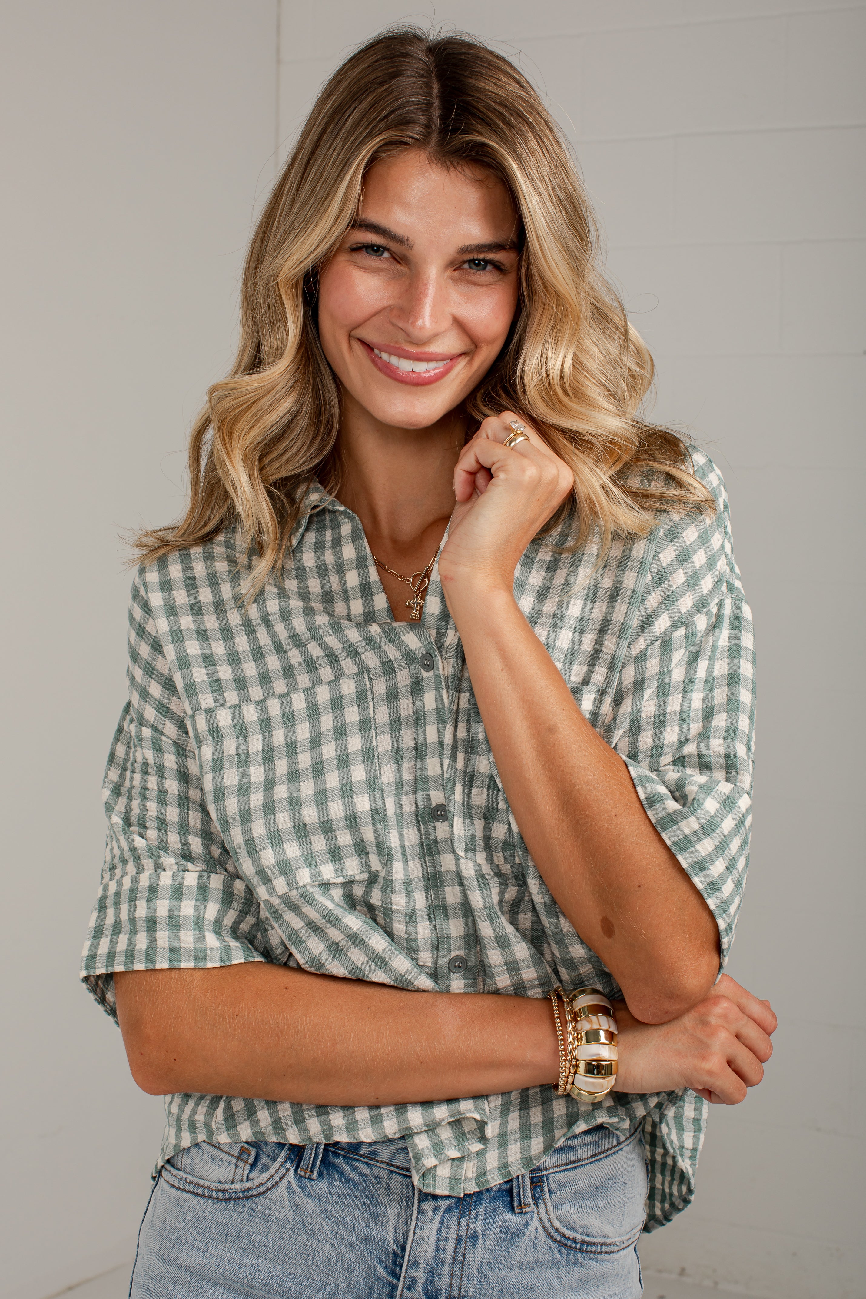 A smiling woman with blonde hair wearing a sage green and white gingham button-down shirt and light-wash blue jeans, posed against a light gray wall.