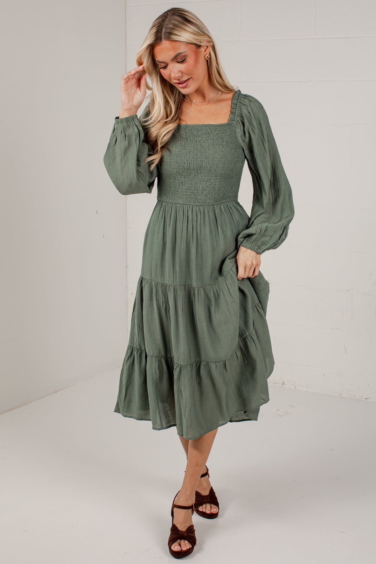 A woman modeling a sage green midi dress with long puff sleeves, a smocked bodice, and a tiered skirt, paired with brown strappy heels against a white studio background.