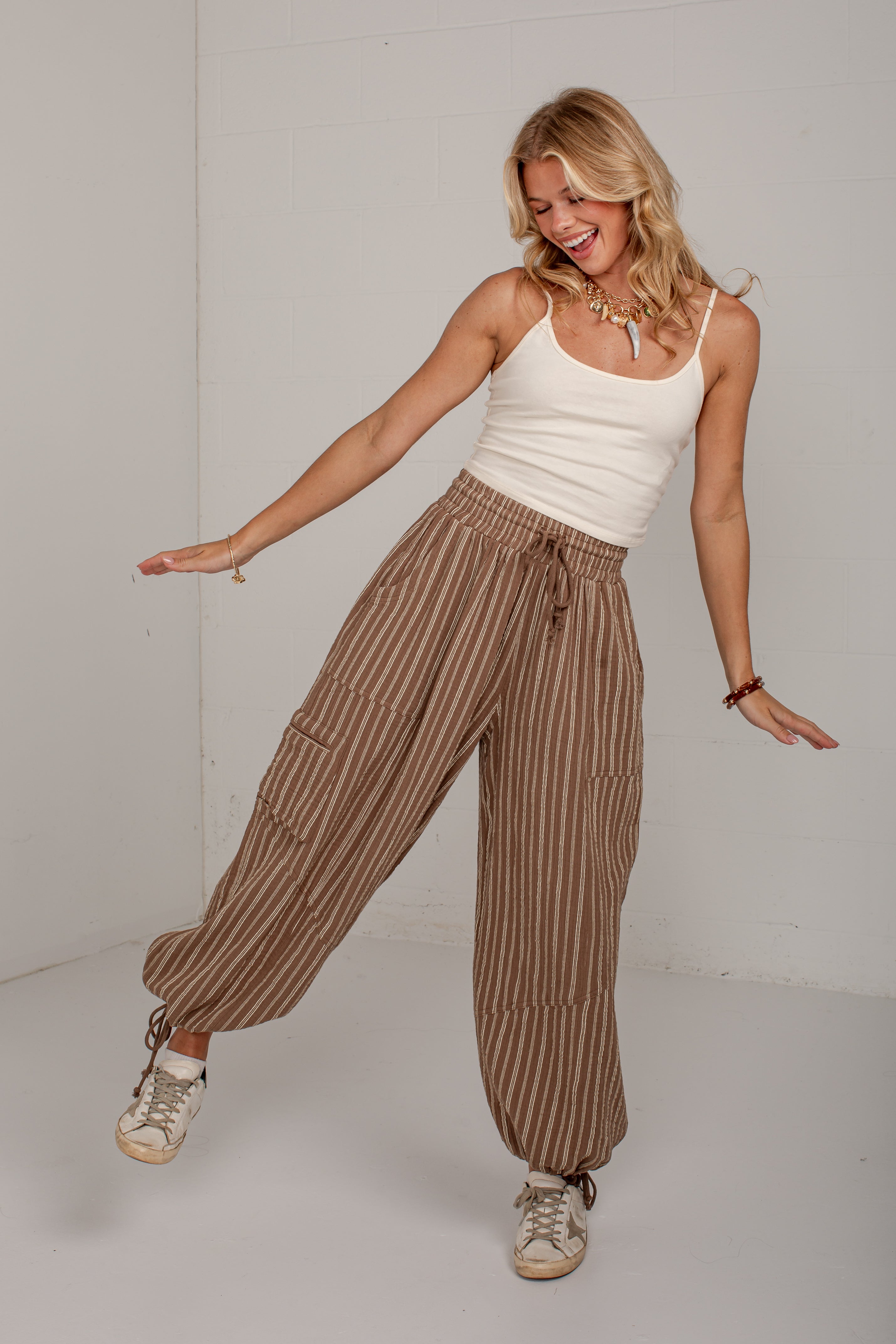 A woman modeling high-waisted, wide-leg striped pants in a studio setting. The pants feature vertical brown and beige stripes, an elastic drawstring waist, and large cargo pockets.
