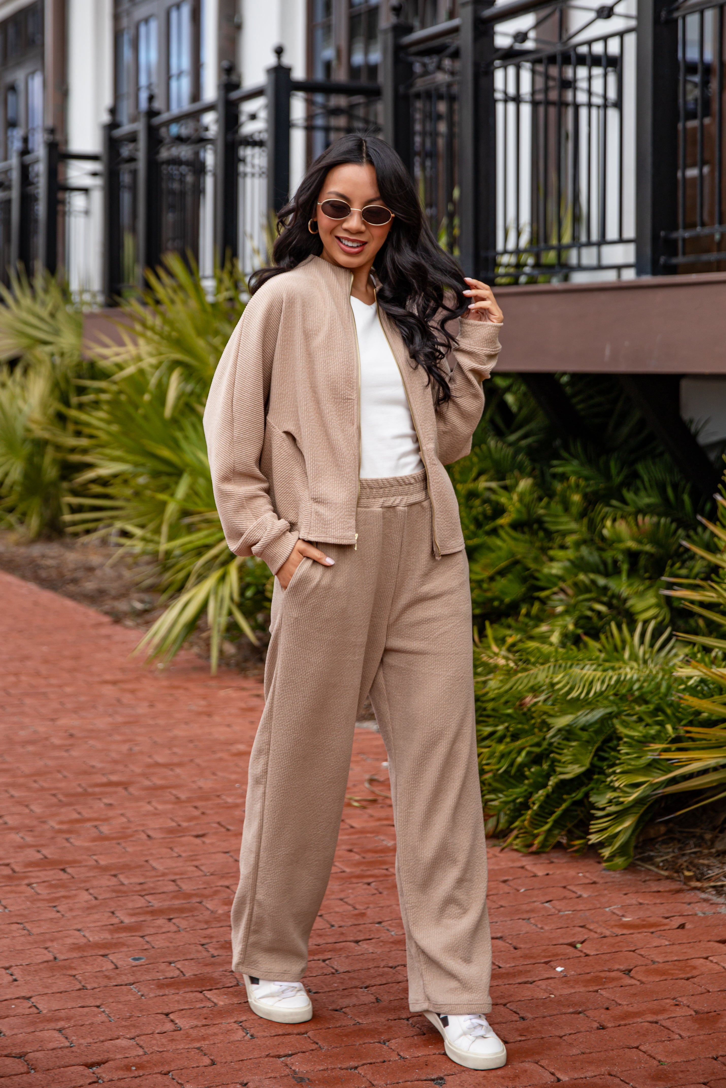 Woman in beige outfit standing on a brick path with greenery in the background