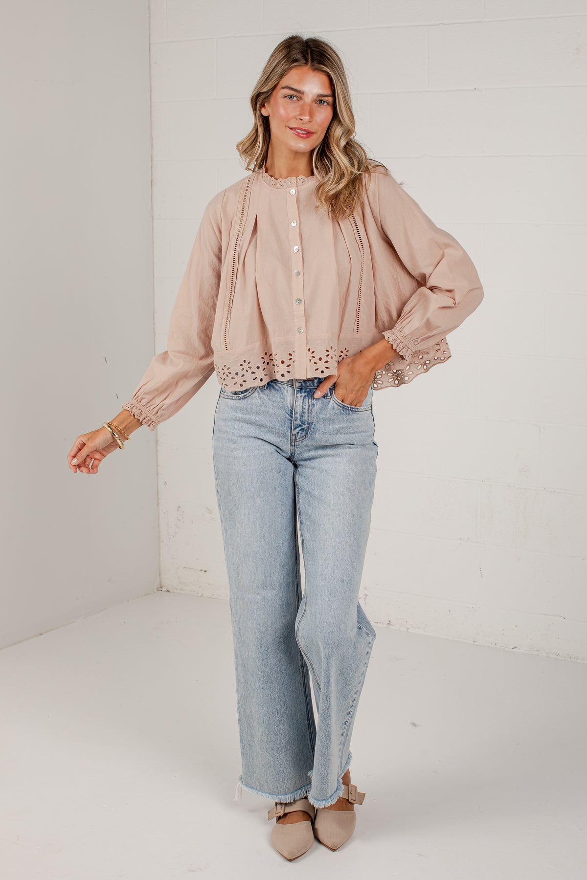 A woman modeling a light blush pink long-sleeve blouse with eyelet lace trim and button details, paired with light-wash wide-leg jeans and beige pointed flats, standing against a white brick wall.