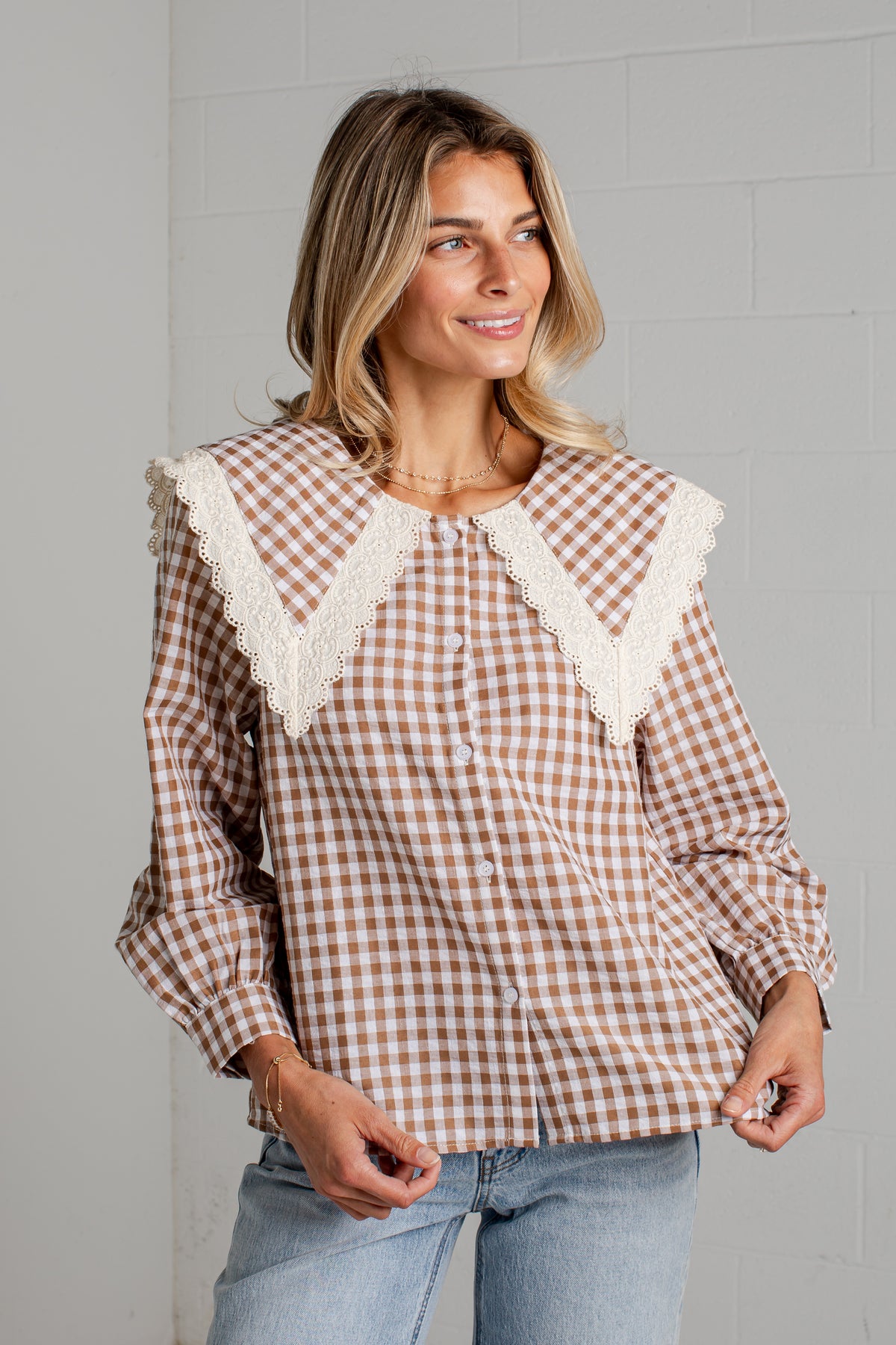 A woman wearing a brown and white gingham long-sleeve blouse with a large cream lace collar, paired with light blue jeans, standing against a light gray brick wall.
