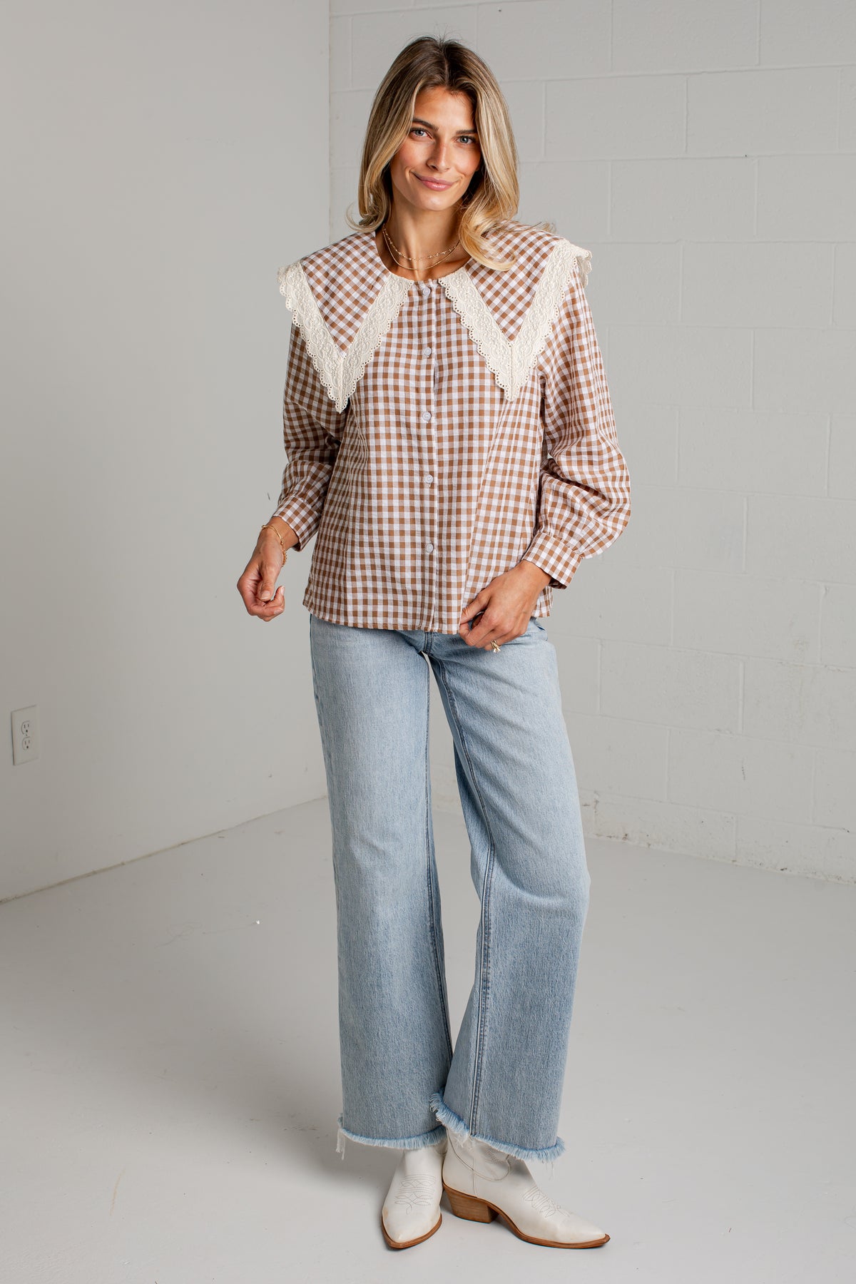 A woman modeling a brown and white gingham long-sleeve blouse with a large cream lace collar, paired with light blue wide-leg jeans and white ankle boots, in a bright studio setting.