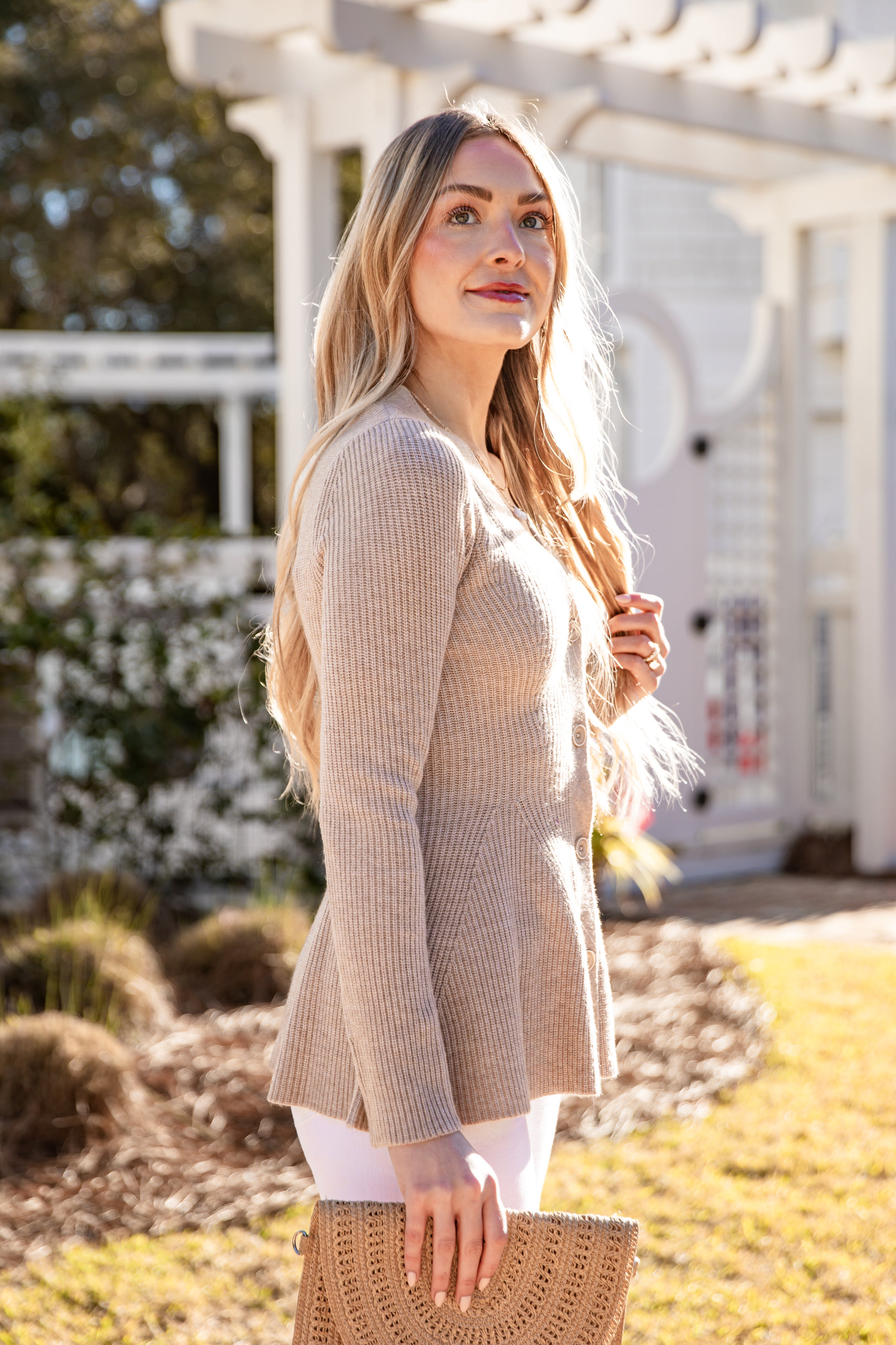 Woman wearing a beige cardigan in an outdoor setting with greenery and a building in the background.