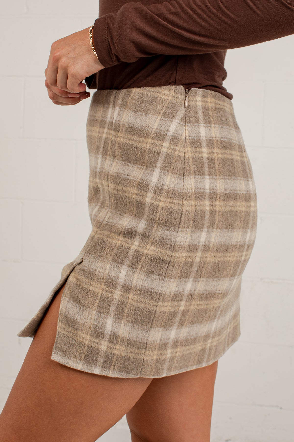 A close-up of a person wearing a brown long-sleeve top and a beige plaid mini skirt with a side slit, standing against a white brick wall.