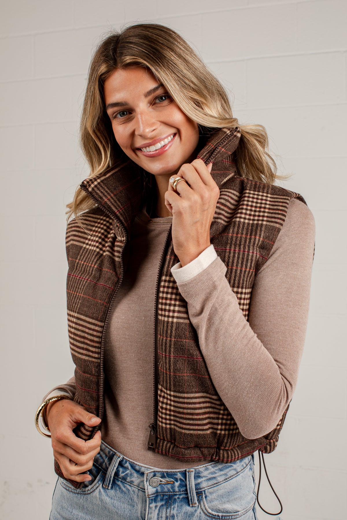 A smiling woman wearing a brown and beige plaid sleeveless puffer vest over a taupe long-sleeve top and blue jeans, standing against a light gray paneled wall.