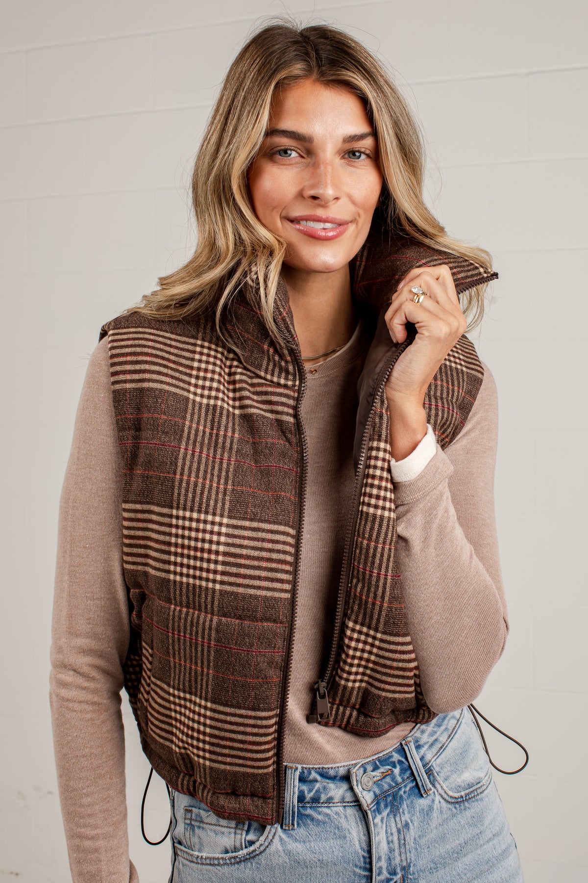 A woman wearing a brown and beige plaid zip-up vest over a beige long-sleeve top and blue jeans, standing against a white paneled wall.