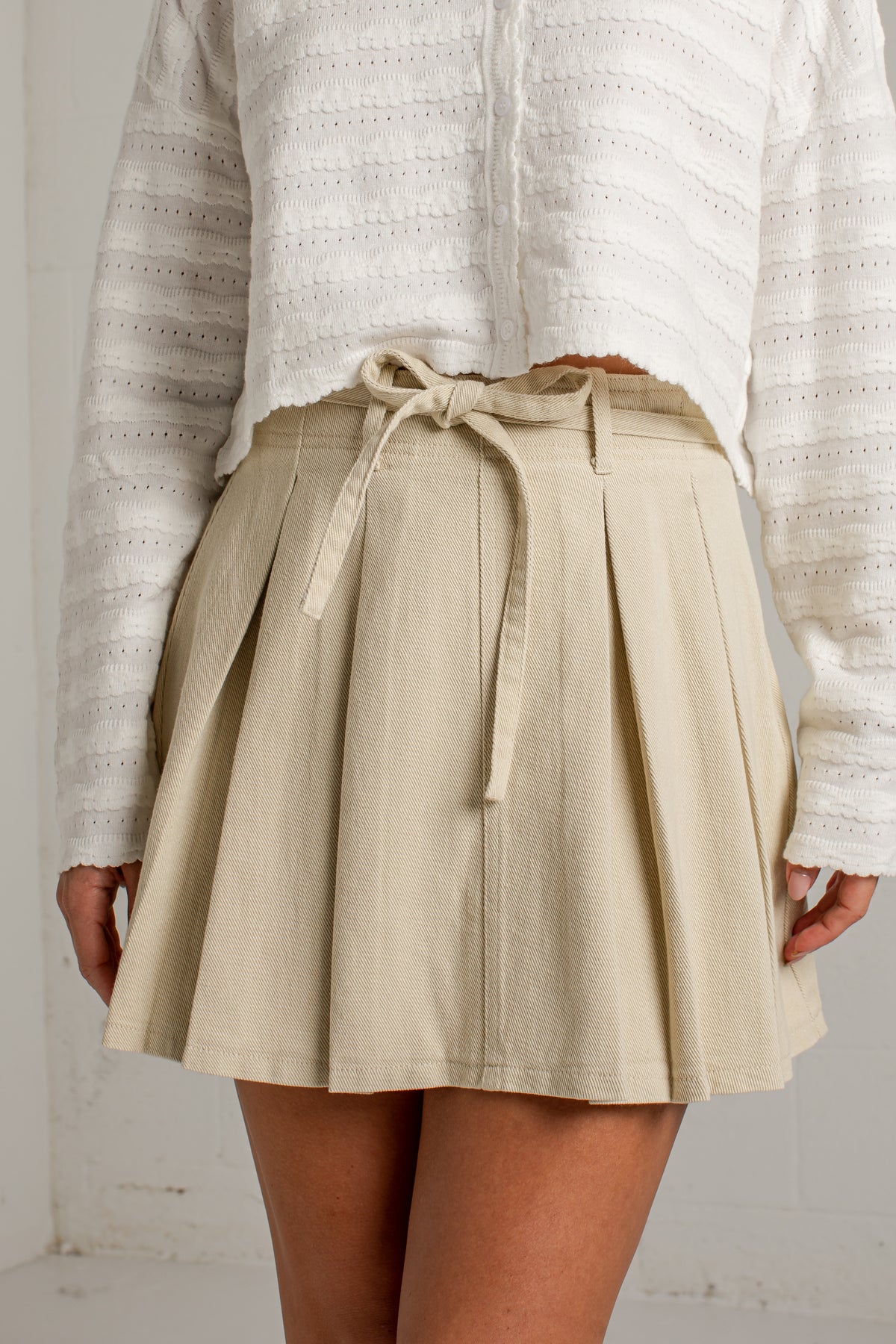 A person wearing a beige pleated mini skirt with a matching fabric tie belt at the waist, paired with a white textured knit top. The photo is cropped from the chest down against a light neutral wall.