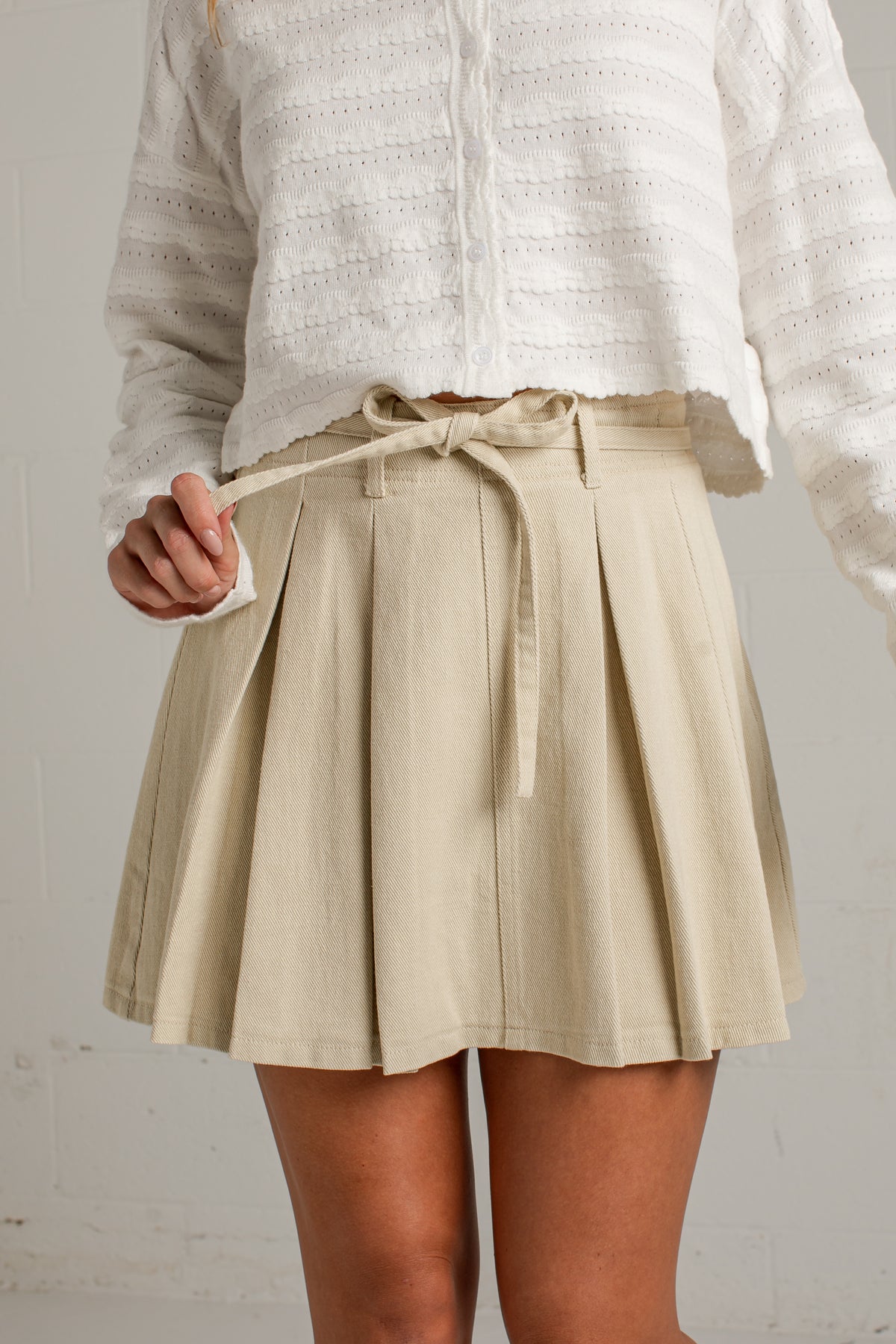 A person wearing a beige pleated mini skirt with a matching tie belt, paired with a white textured knit top, shown from the chest down against a light neutral wall.