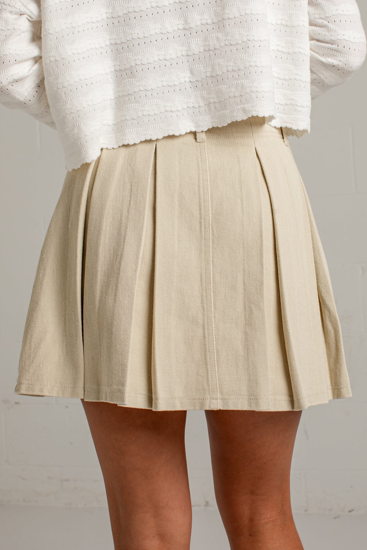 A person wearing a beige pleated mini skirt and a white textured knit top, shown from the chest down against a light neutral wall.