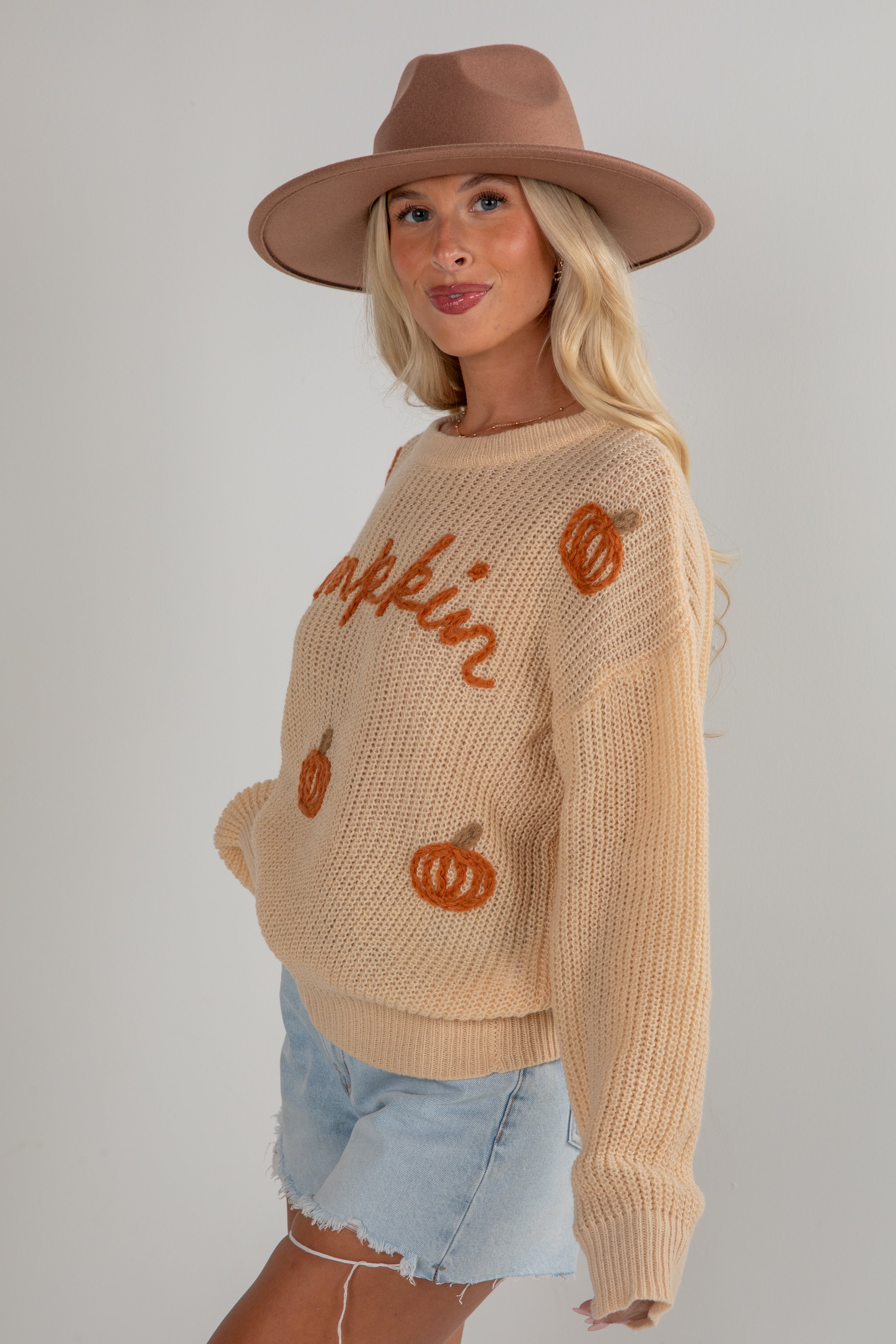 Woman wearing a beige sweater with pumpkin graphics and a brown hat on a plain background
