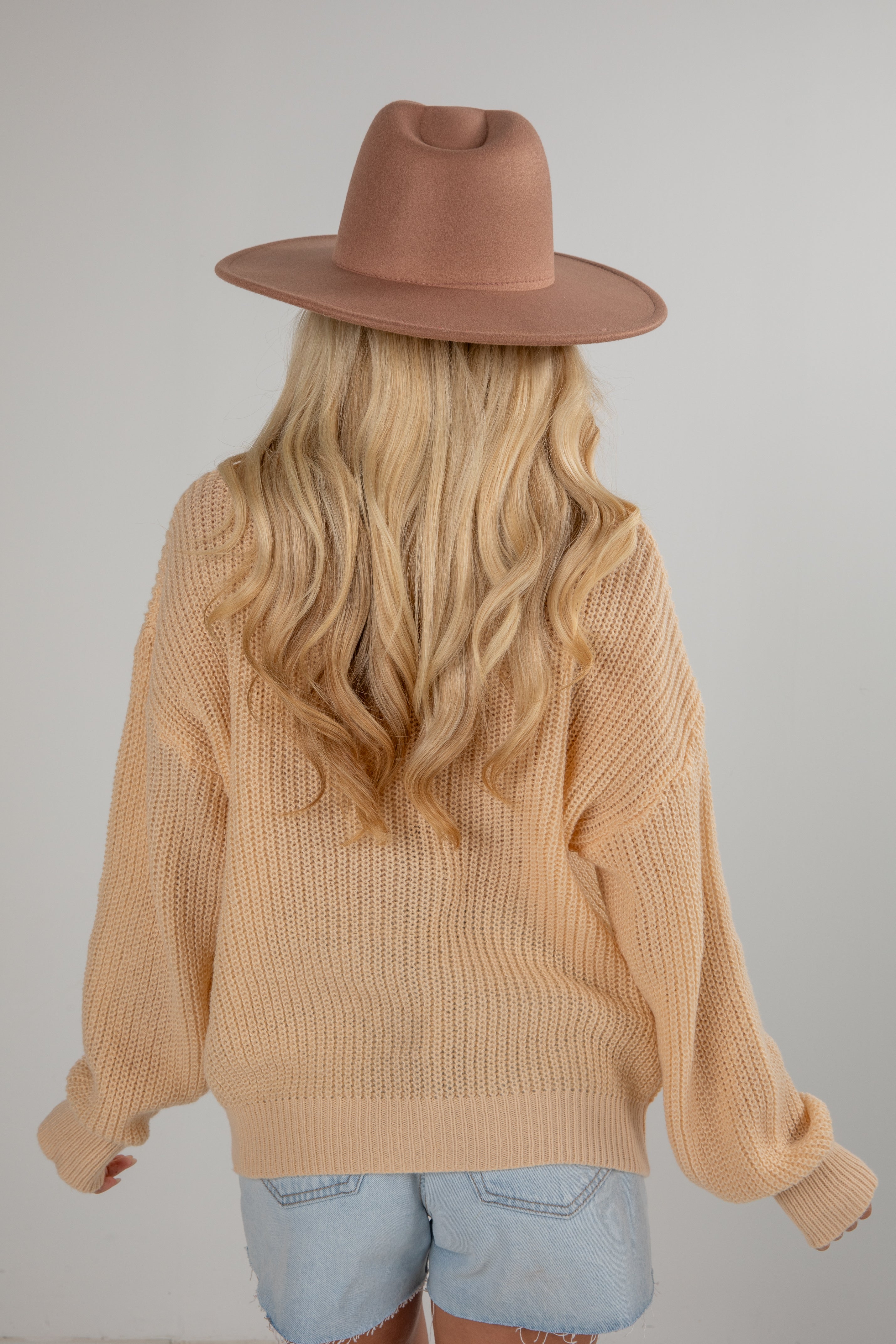 Person wearing a beige sweater and brown hat with long blonde hair, standing against a plain background