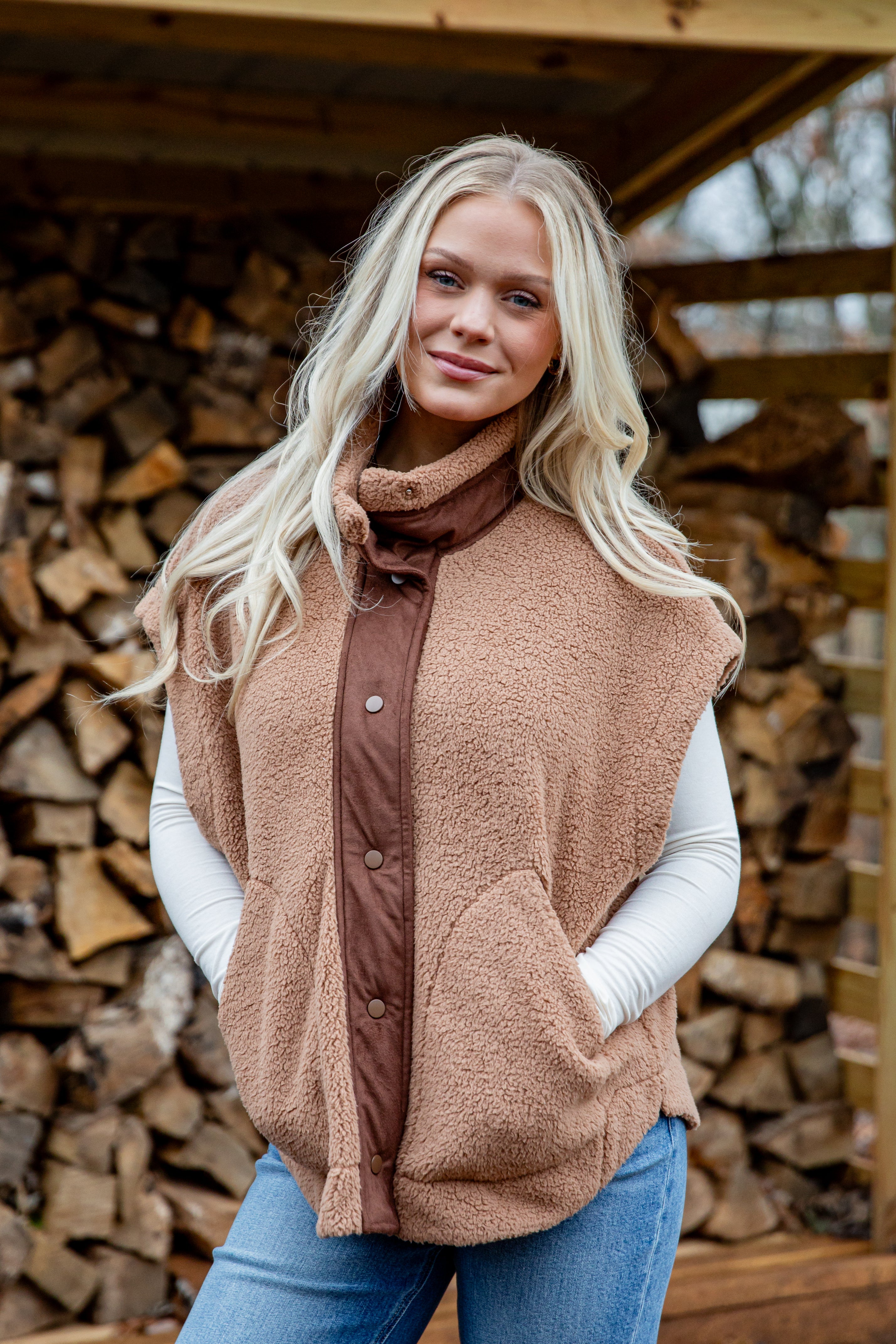 Woman wearing a brown quilted vest over a white shirt and blue jeans, standing in front of stacked firewood.