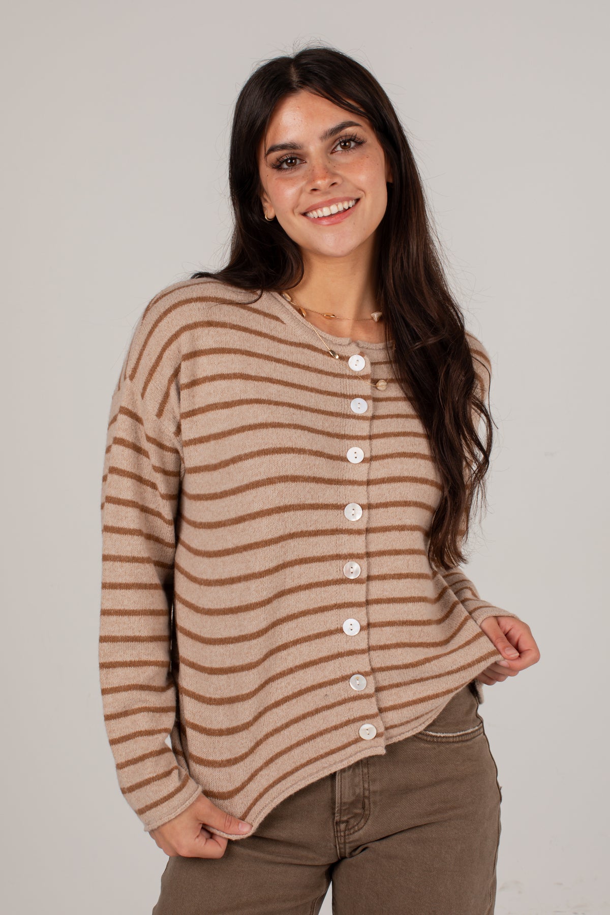 A woman with long dark hair smiling while wearing a beige and brown horizontally striped button-up cardigan sweater and brown pants, photographed against a plain light gray studio background.