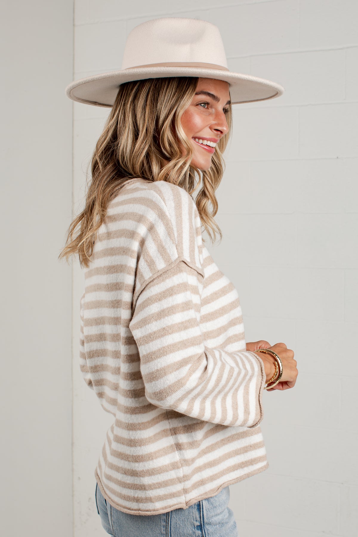 A woman wearing a cream and beige horizontally striped knit sweater, a wide-brimmed cream hat, and light blue jeans, standing against a white paneled wall.