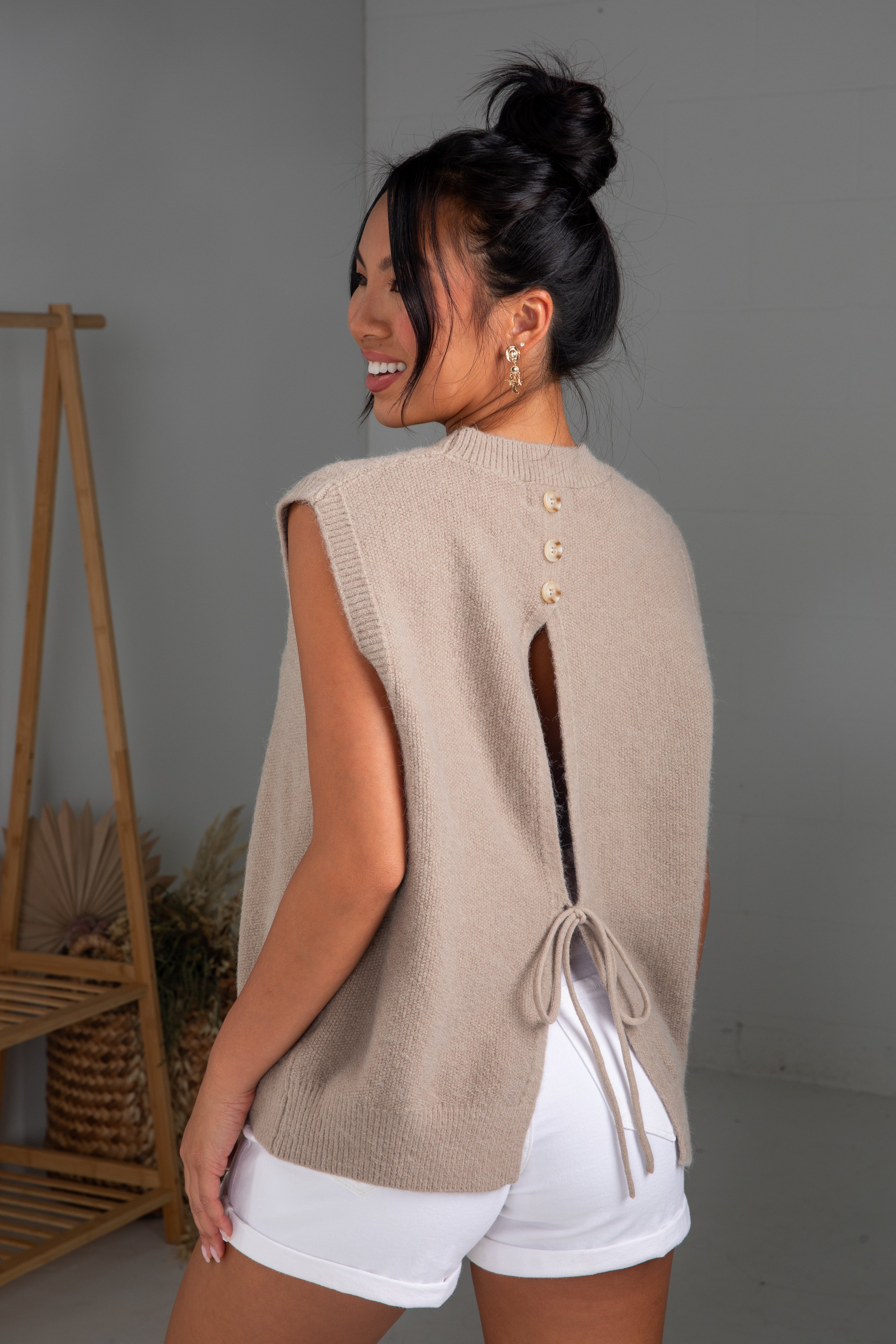 Woman wearing a beige sleeveless sweater with button details and white shorts, standing against a plain background.