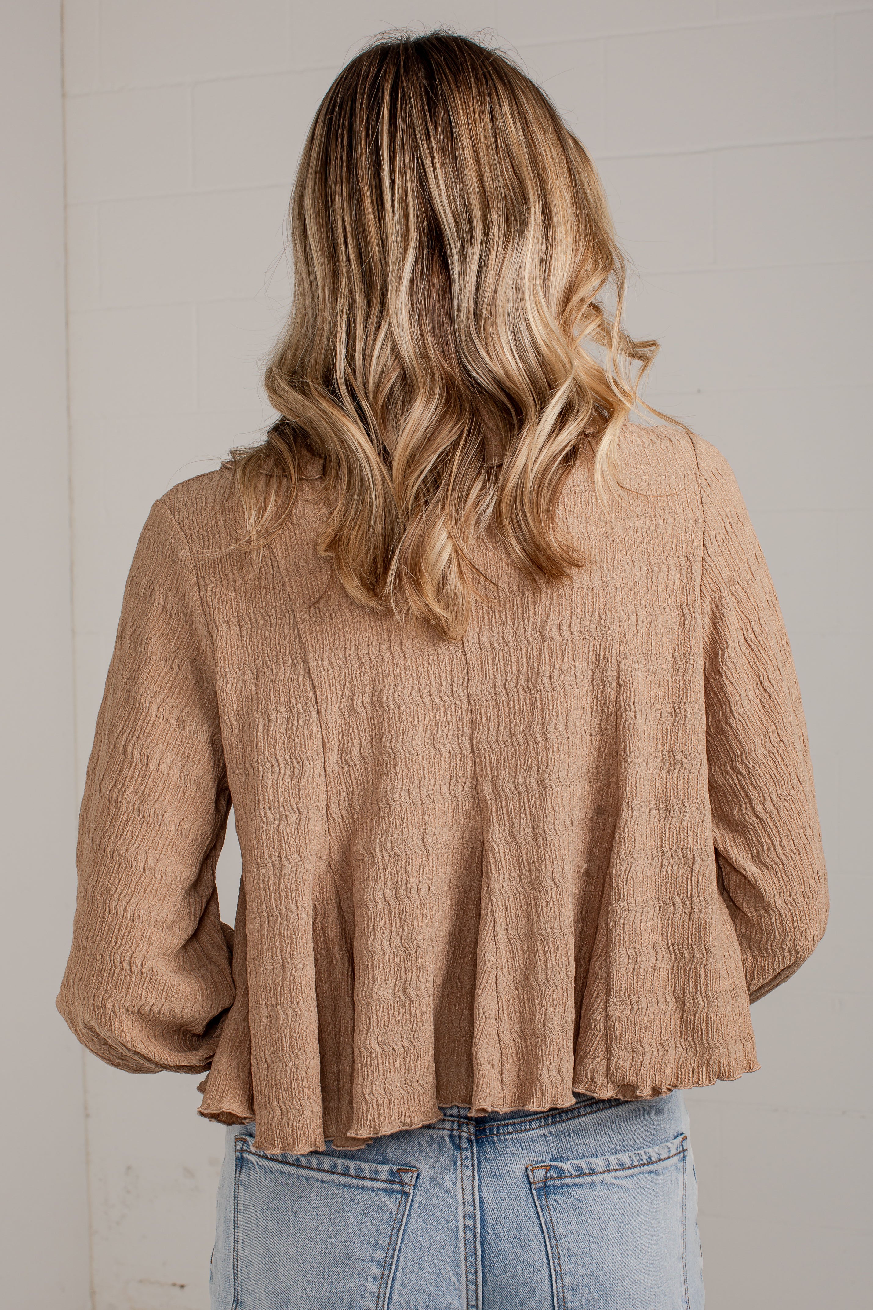 A woman viewed from behind wearing a textured, light brown crinkle blouse and light blue jeans against a white paneled wall.