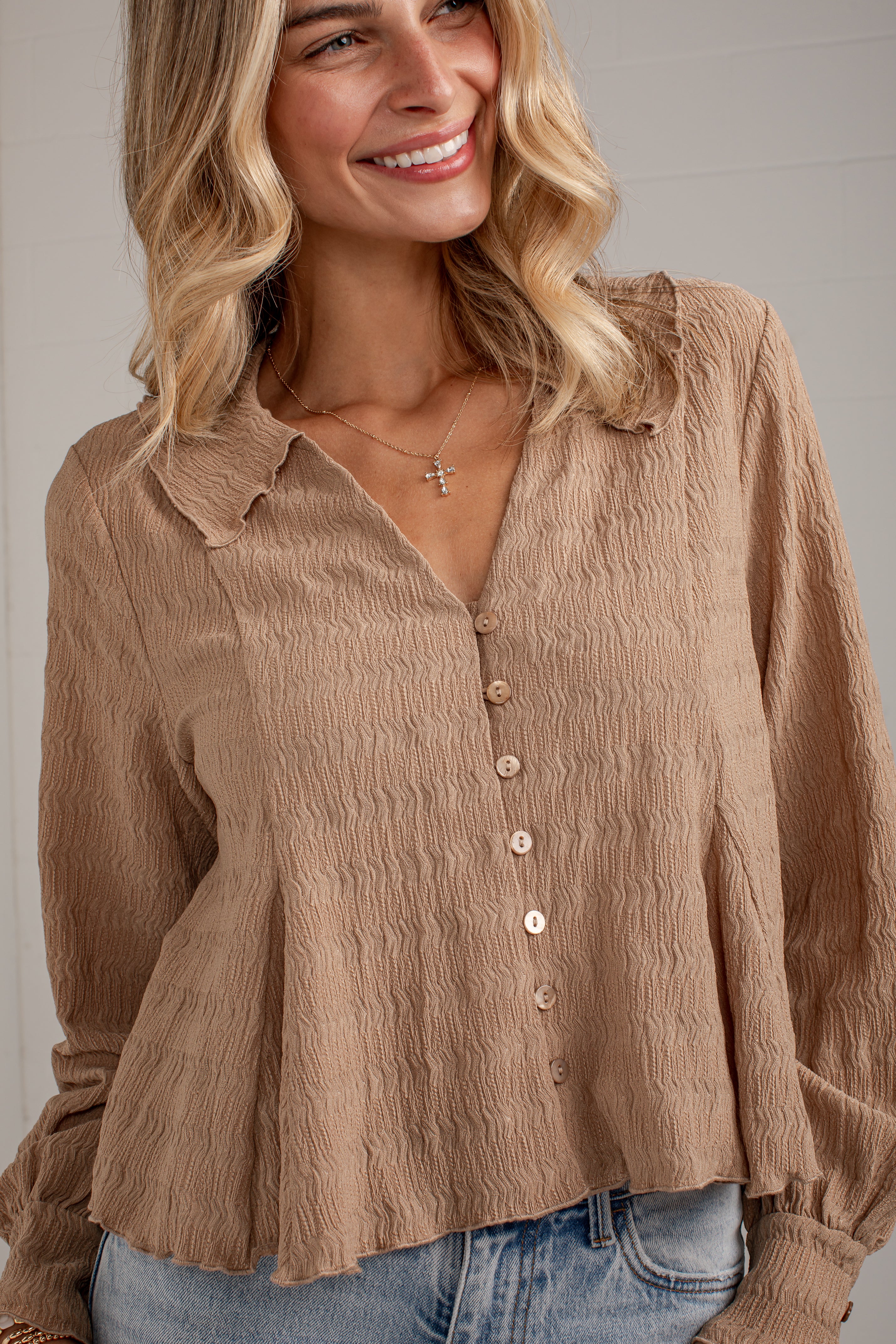 A woman wearing a crinkled taupe-brown long-sleeve button-up blouse with a collar and light blue jeans, shown from the waist up against a neutral indoor background.