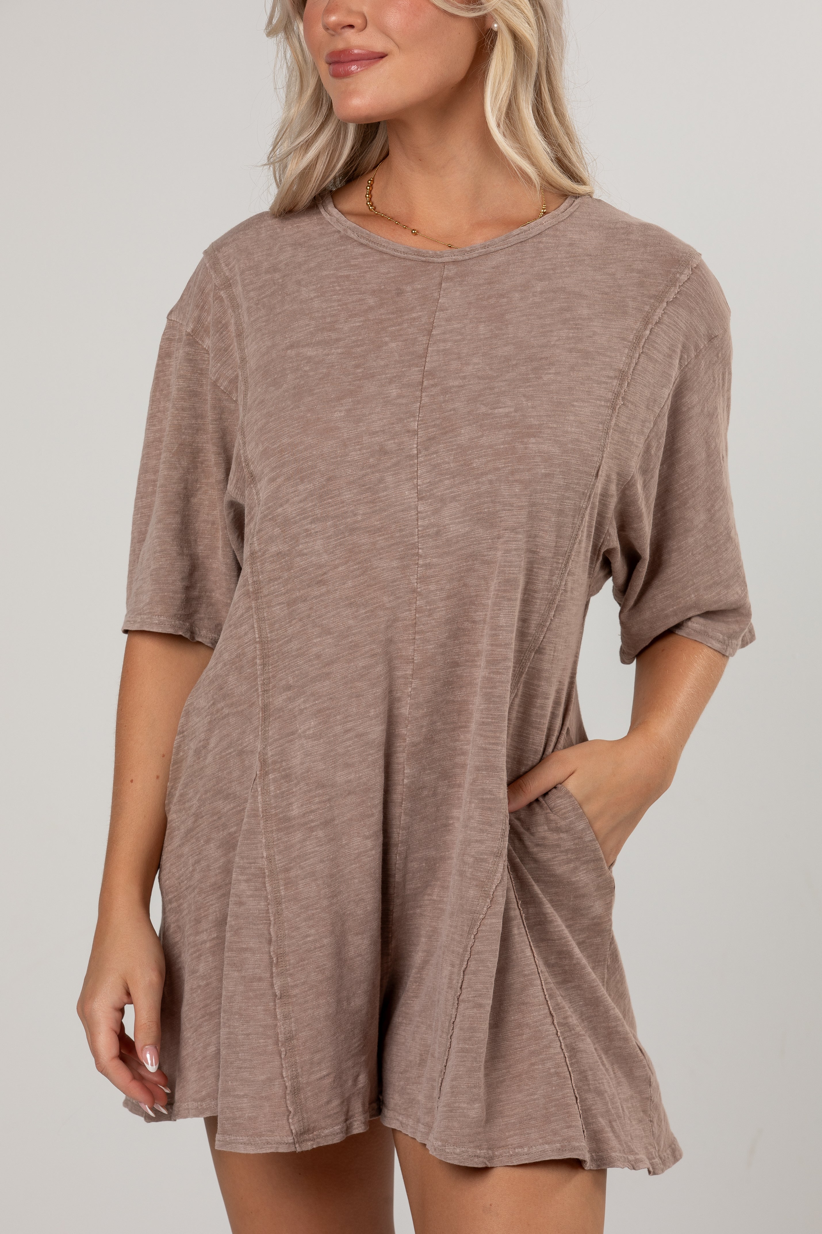 A woman wearing a loose, short-sleeve taupe-brown romper with side pockets, shown from the neck down against a light gray studio background.