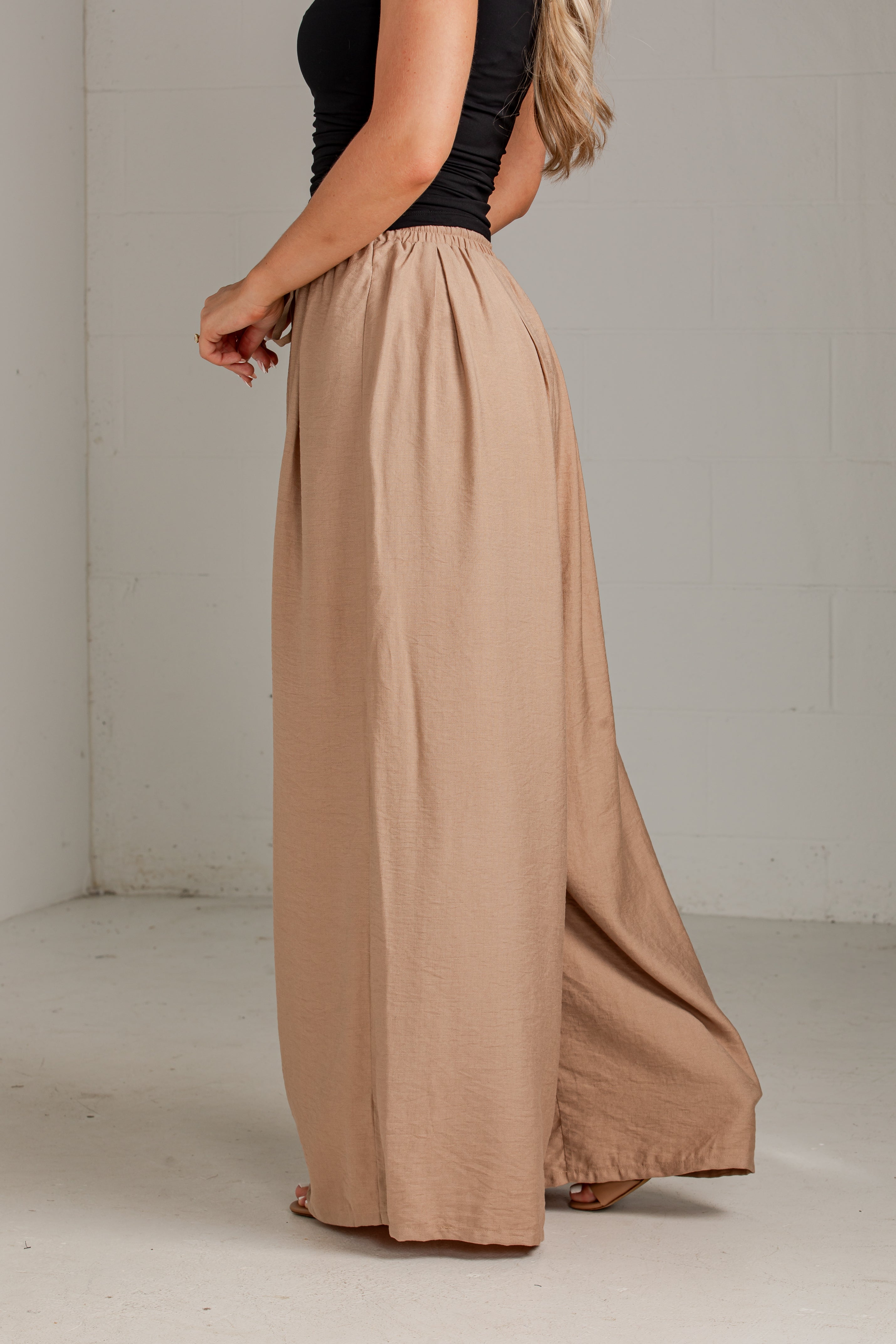 A woman wearing a black sleeveless top and high-waisted, wide-leg taupe maxi pants, standing in a minimalist studio with a white brick wall background.