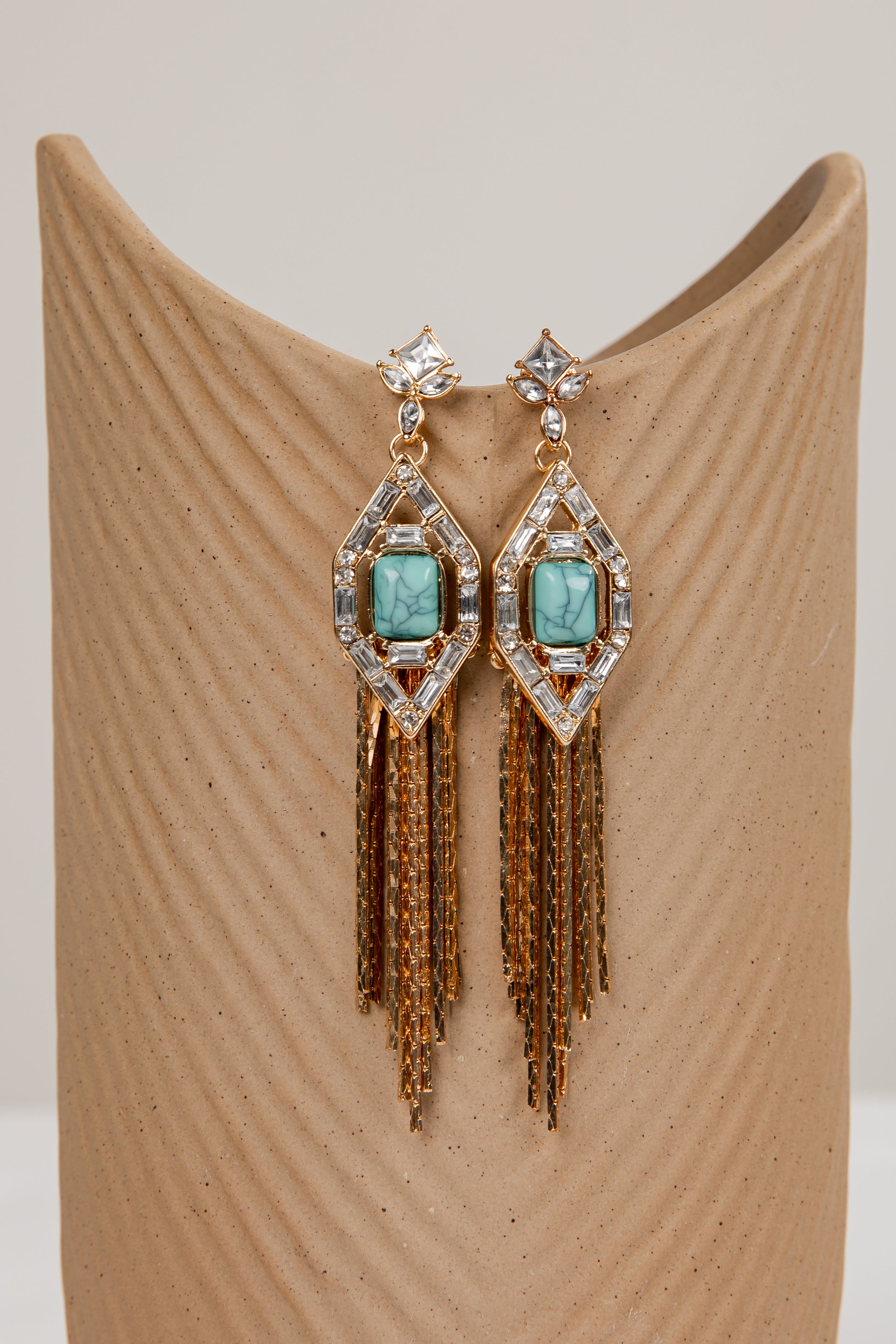 Pair of earrings with turquoise stones and gold fringes on a beige stand.