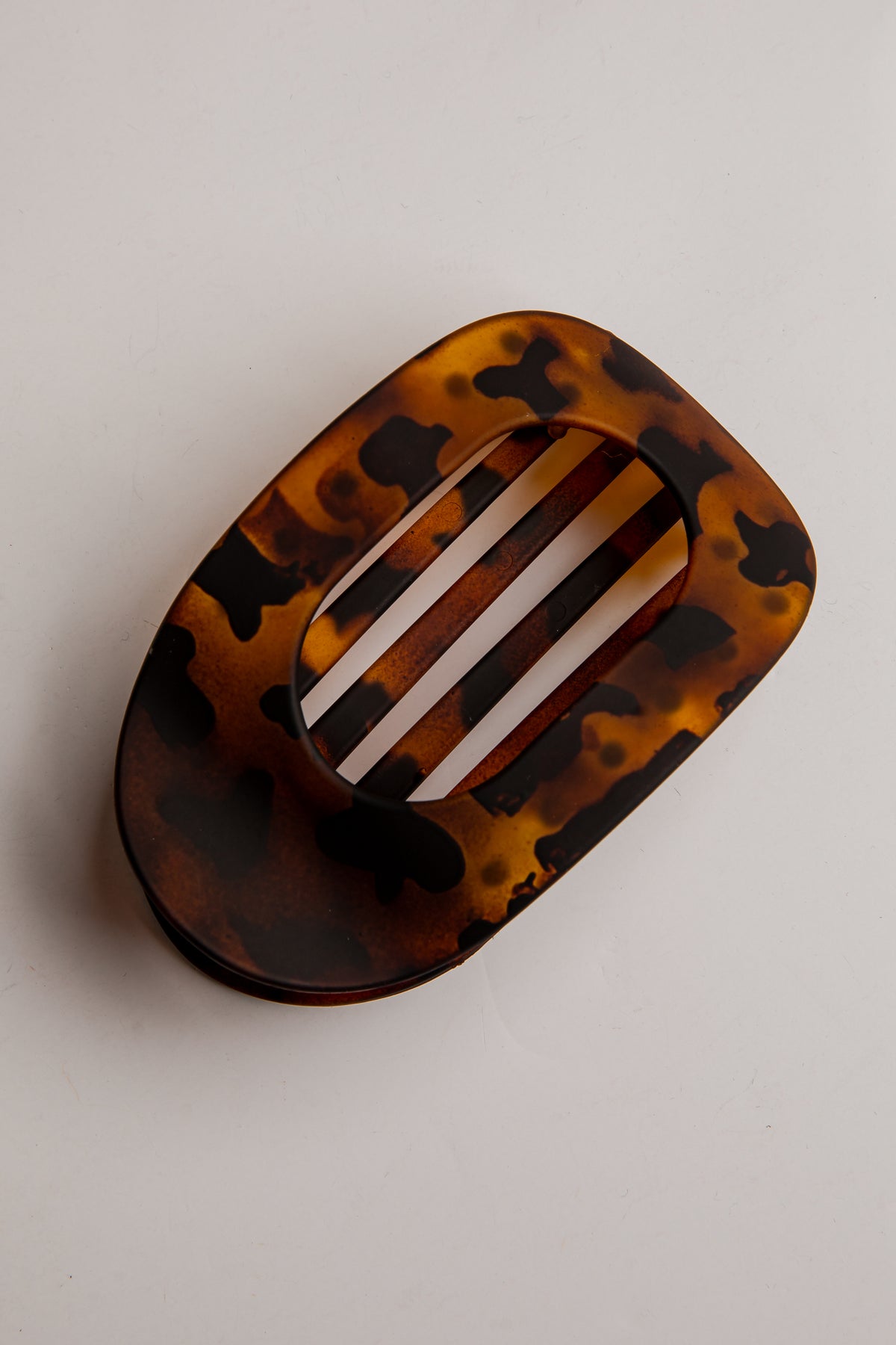 A tortoiseshell hair claw clip with a glossy amber, brown, and black pattern, featuring a central cutout with three white stripes, shown on a neutral light gray background.
