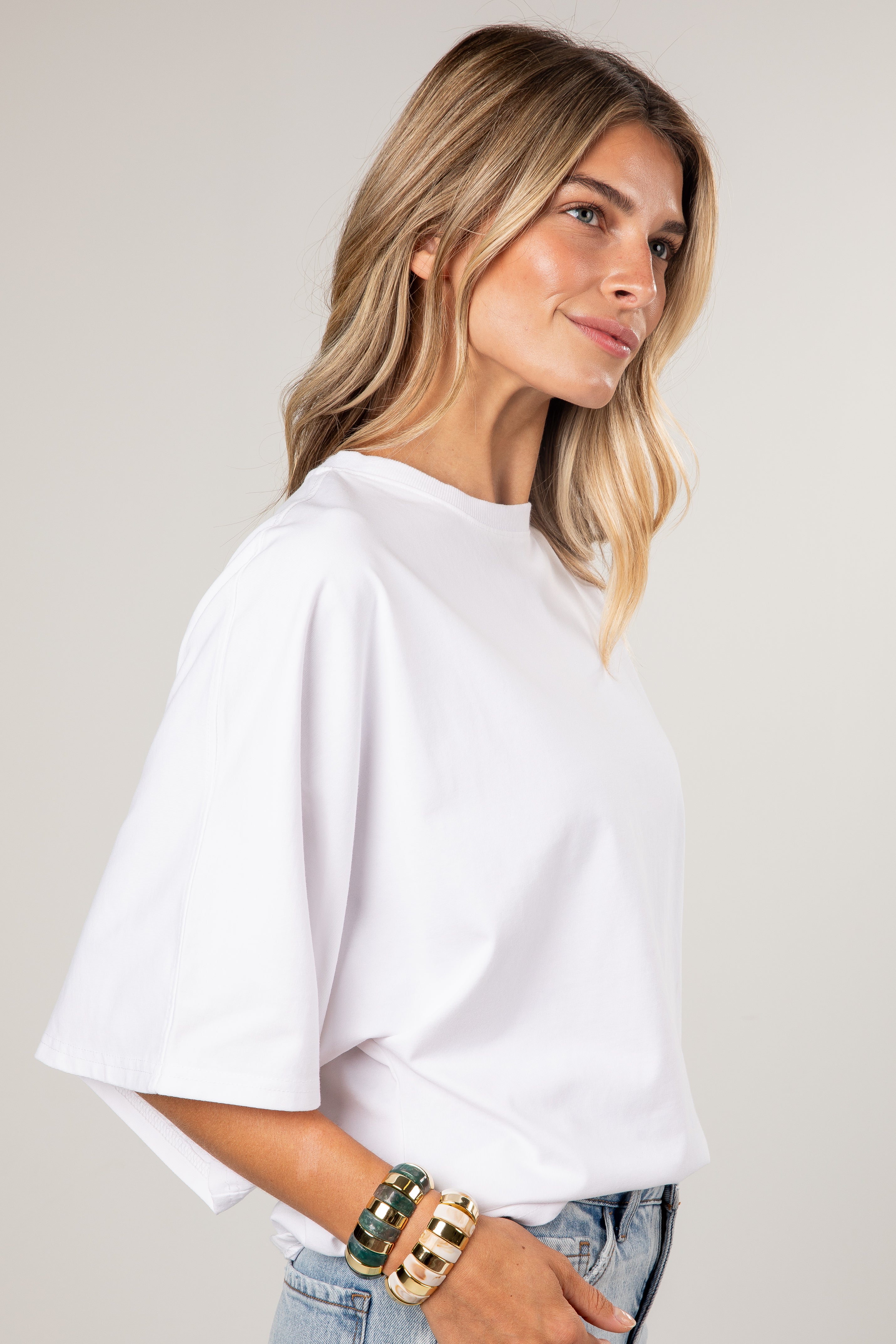 A woman wearing a white short-sleeve t-shirt with a relaxed, oversized fit and a crew neckline, paired with light-wash blue jeans and gold bracelets, set against a plain light gray background.