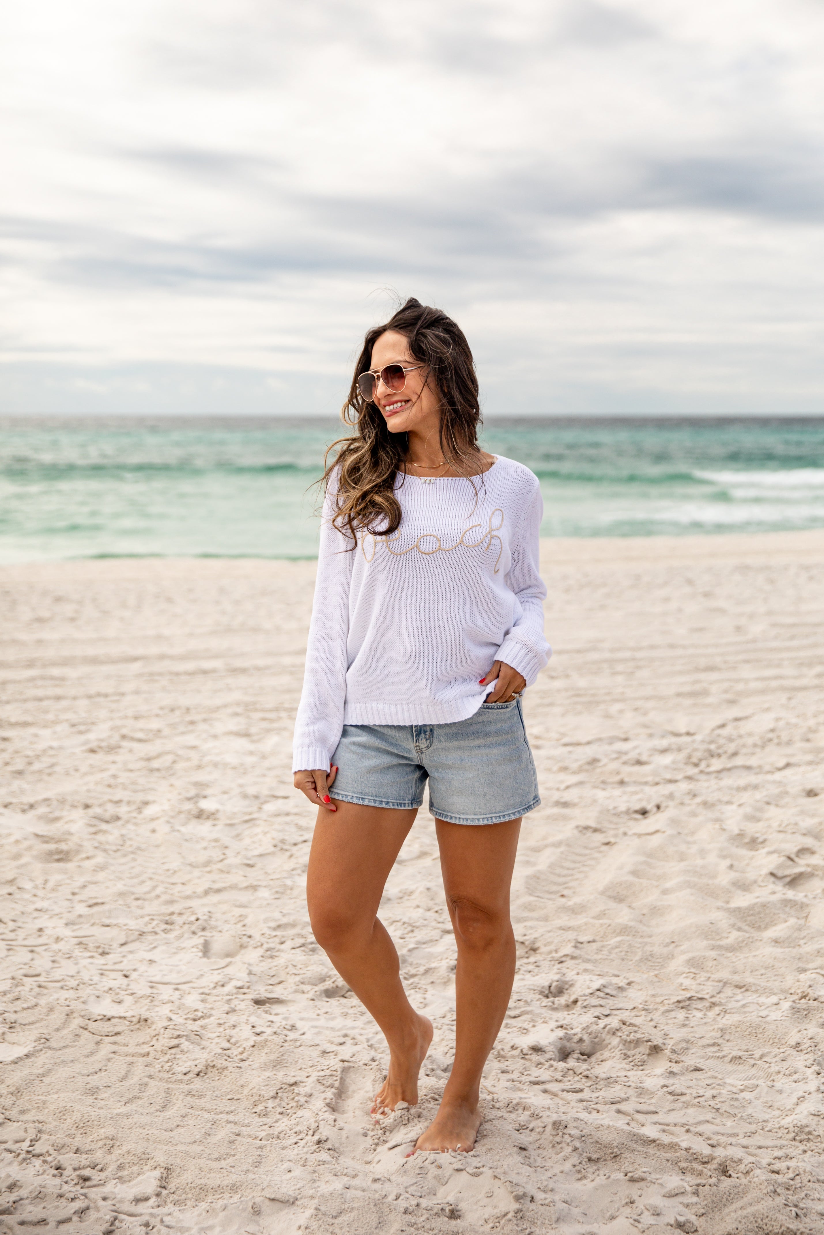 Woman standing on a beach wearing a white long-sleeve shirt and denim shorts.